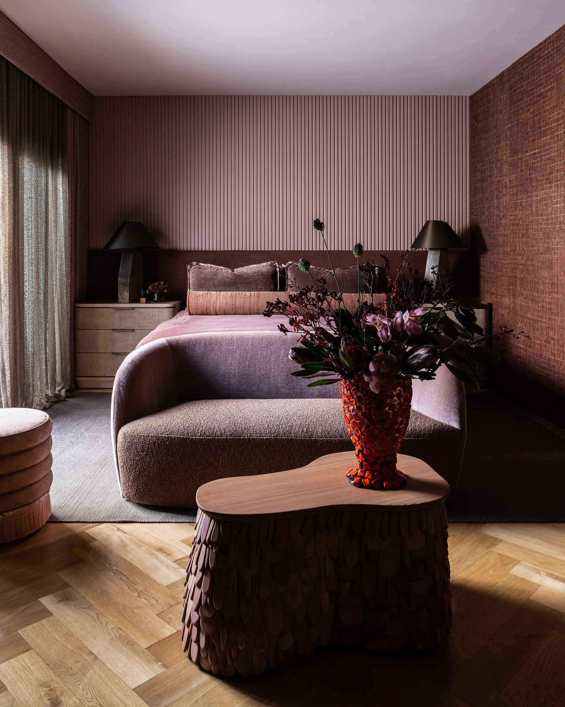 A contemporary bedroom features a curved mauve bench and textured wood coffee table on a herringbone floor in front of a bed with a ribbed headboard wall.