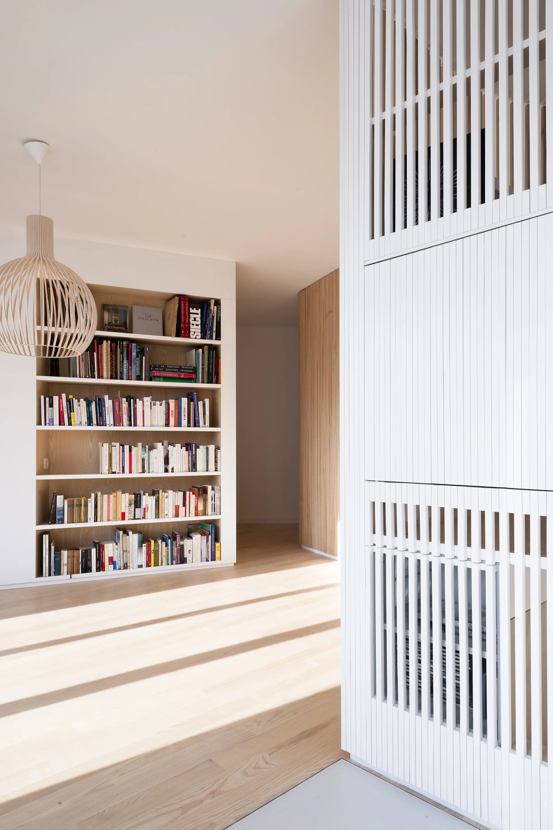 A minimalist home library features a recessed wood bookshelf filled with books, a white slatted room divider in the foreground, and light wood flooring.