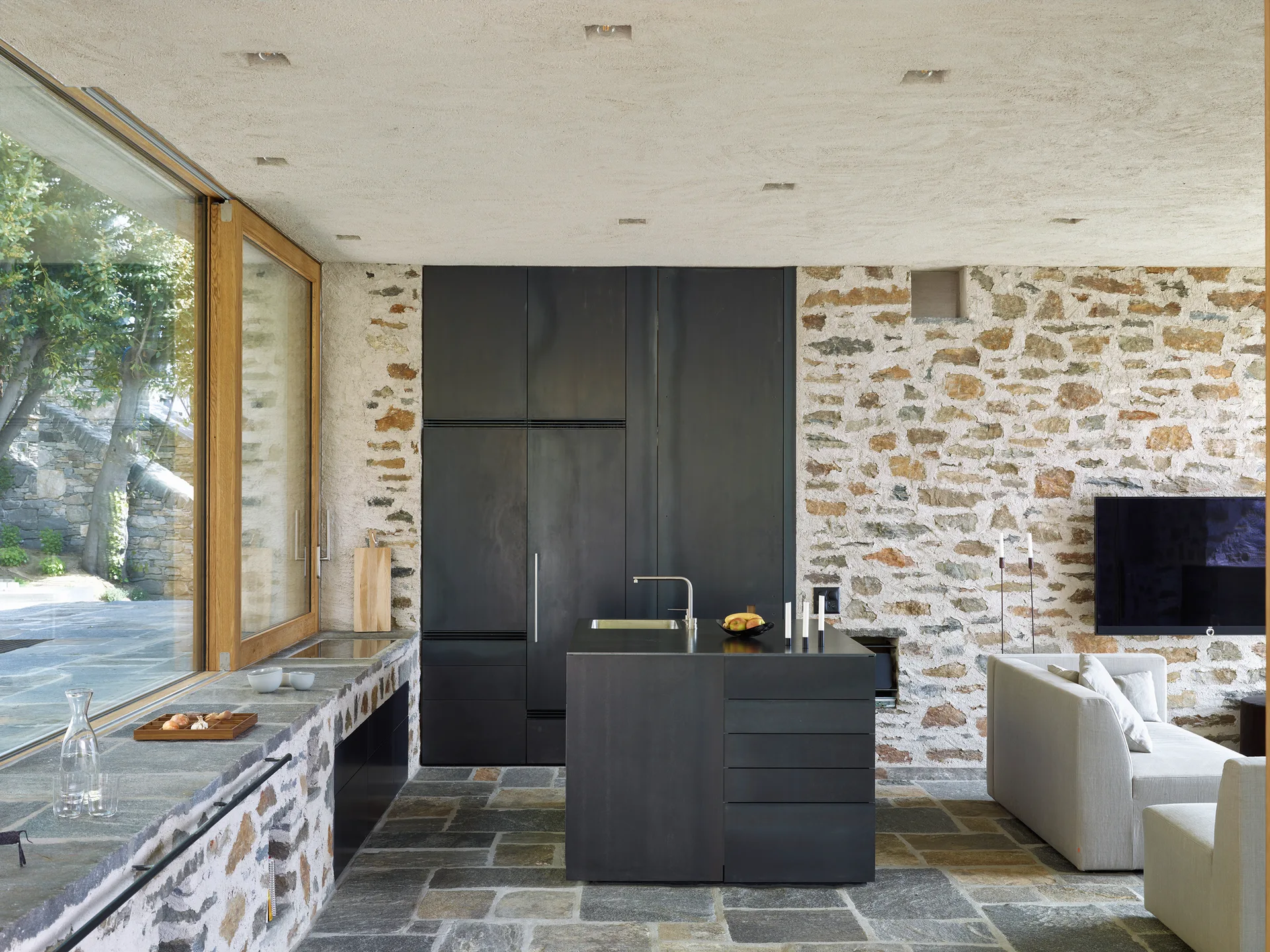 A minimalist kitchen featuring a dark matte island and tall cabinetry set against a rough stone wall and irregular stone floor tiles.