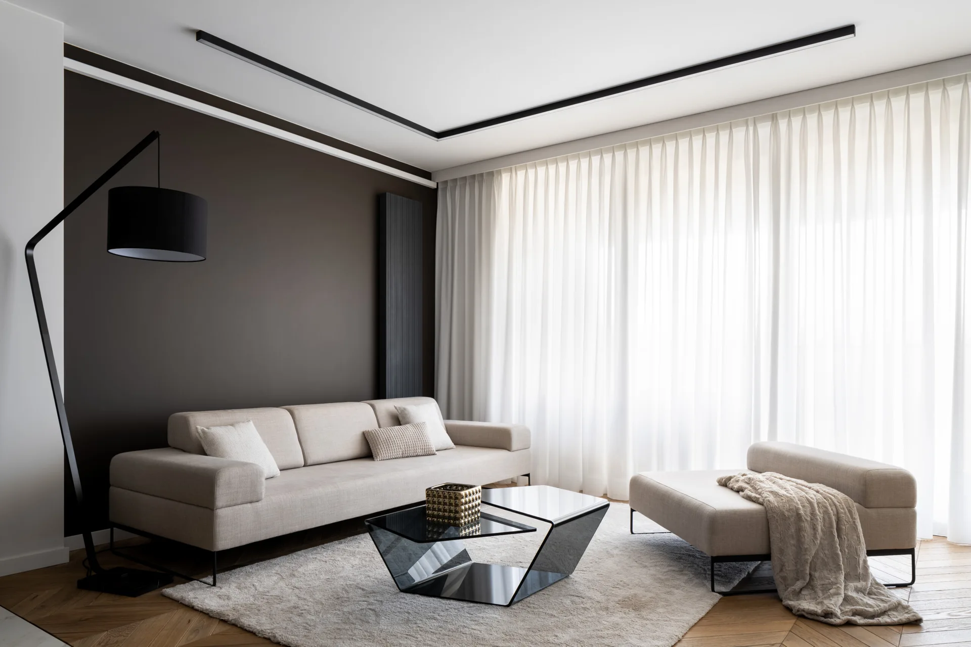 A contemporary living room featuring a beige sofa, a geometric glass coffee table, a dark brown accent wall, and sheer white curtains.