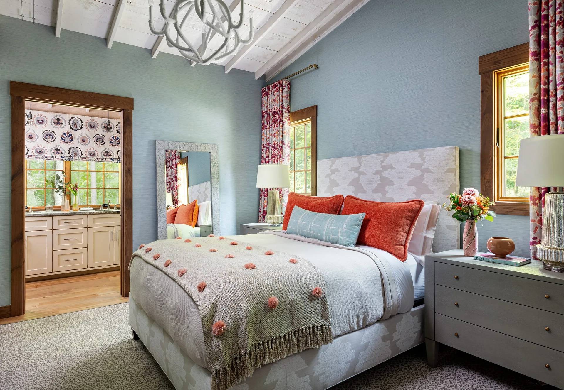 A bedroom featuring an upholstered bed with orange pillows, light blue wallpaper, patterned curtains, and a vaulted ceiling with white beams.