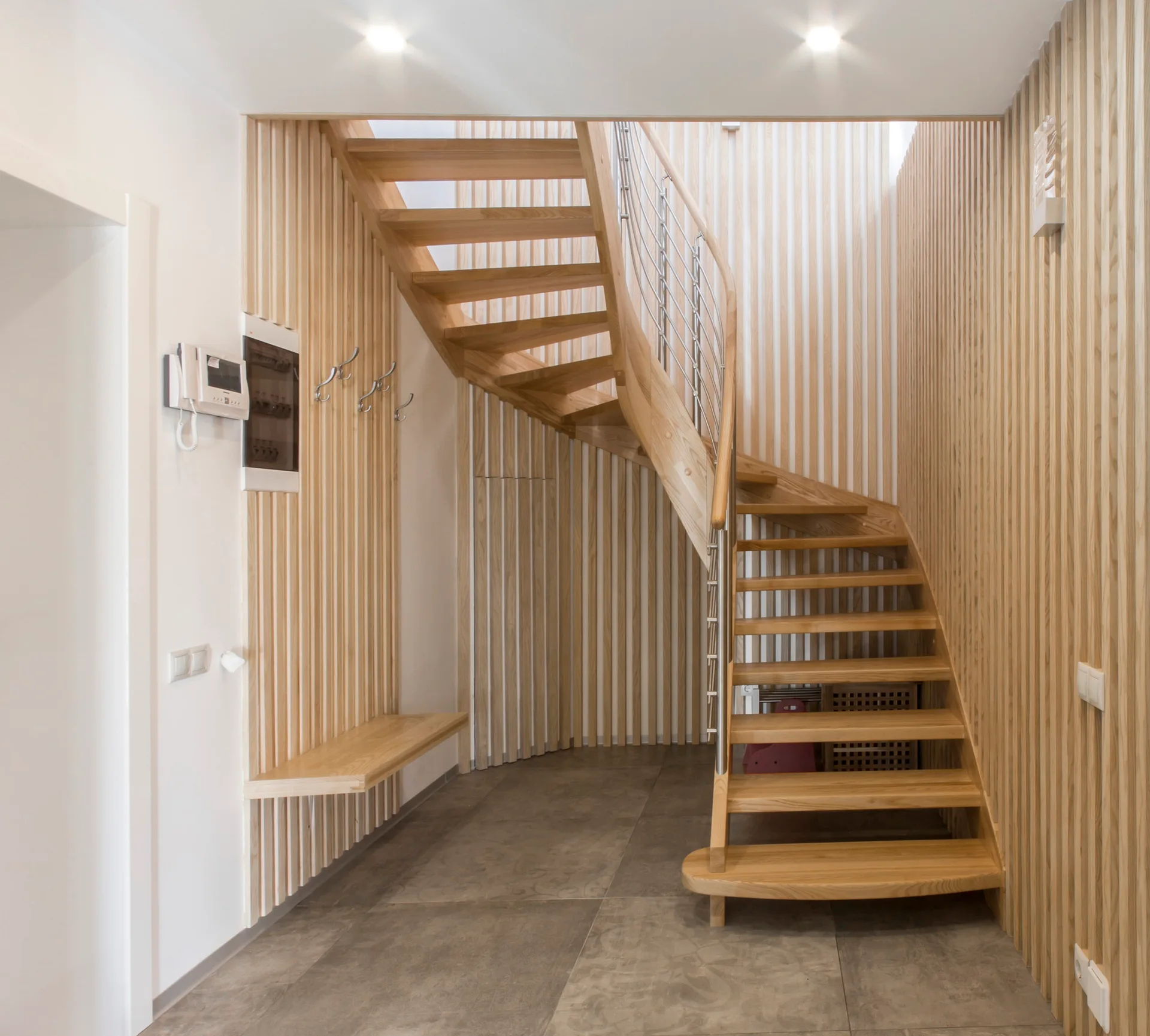 A curved wooden staircase rises against walls clad in vertical timber slats next to a floating wooden bench on grey floor tiles.