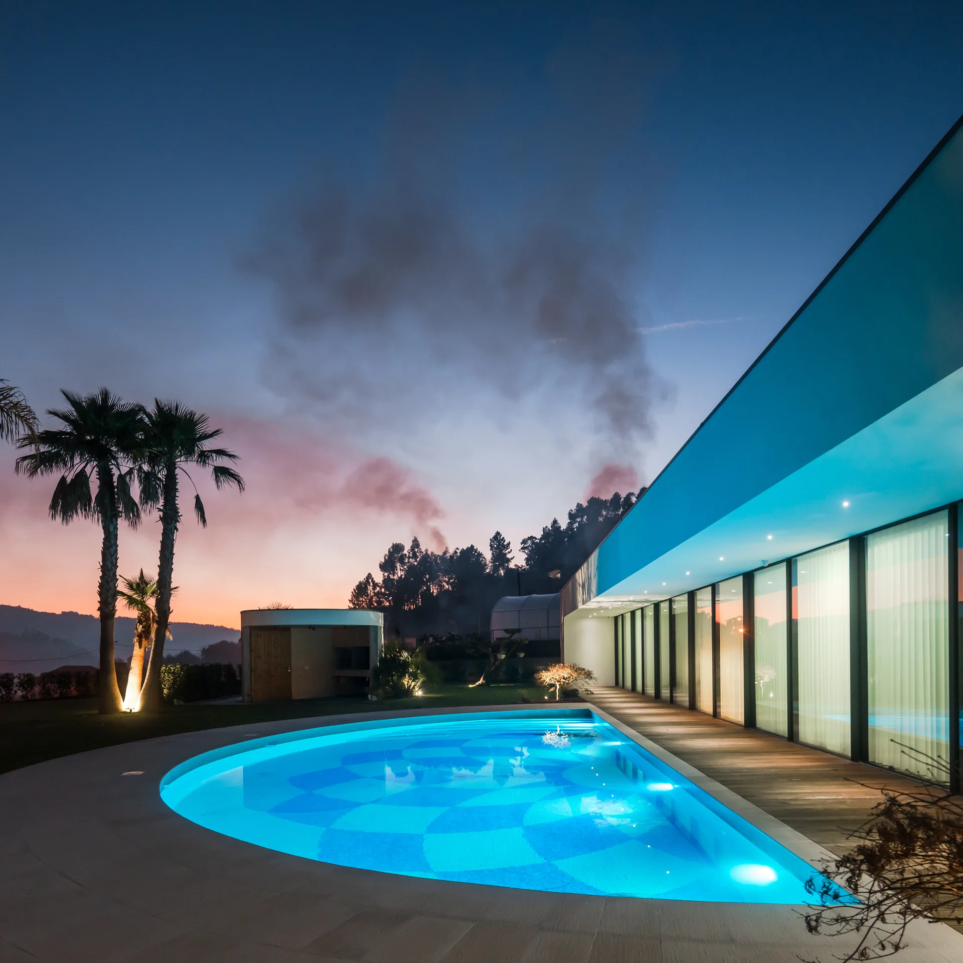 A modern house with glass walls features a curved, illuminated blue swimming pool set beside a wood plank deck and palm trees at twilight.