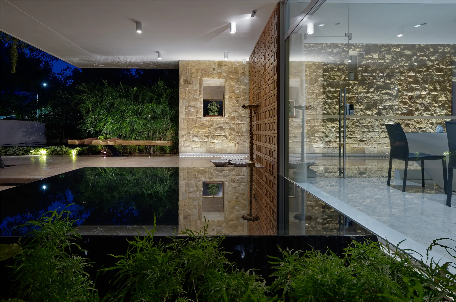 A modern covered patio featuring a dark reflecting pool next to a textured stone wall, wooden lattice panel, and glass entry.
