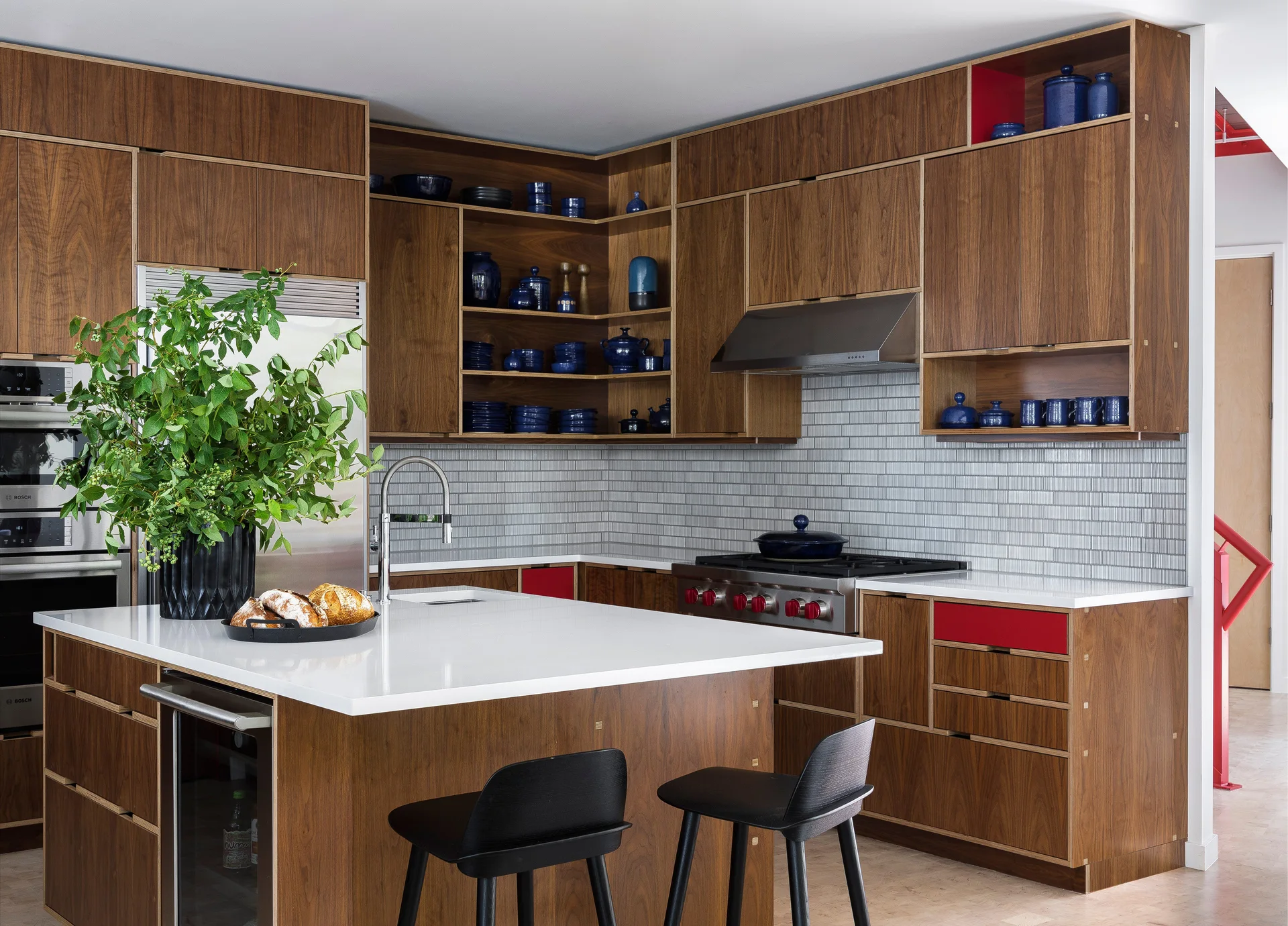 A modern kitchen with warm wood cabinetry, a grey tiled backsplash, a white-topped island with black stools, and blue ceramic dishes on open shelves.