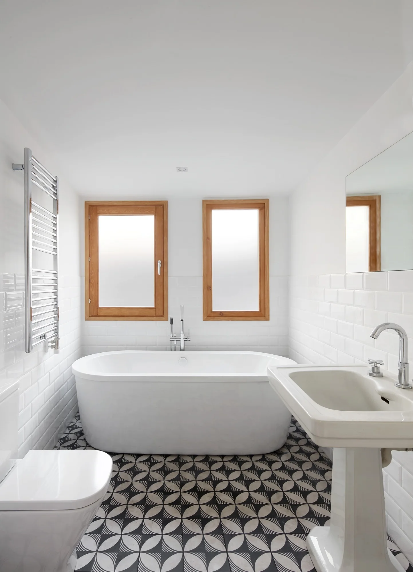 A bright bathroom with a white freestanding bathtub positioned between two wood-framed windows, a pedestal sink, and geometric floor tiles.