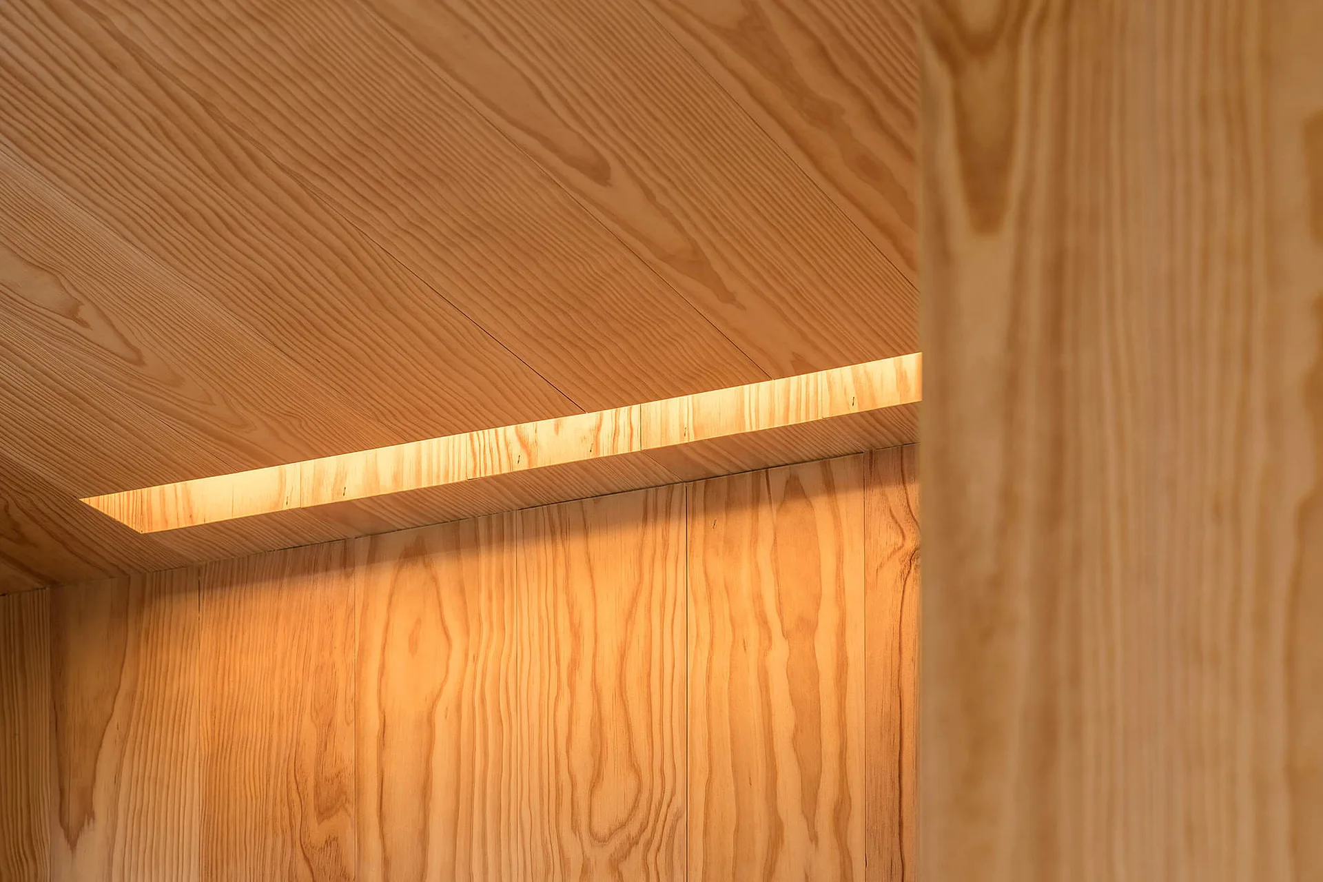 Warm recessed cove lighting glows from a narrow niche where a sloped wood paneled ceiling meets a matching wood wall.