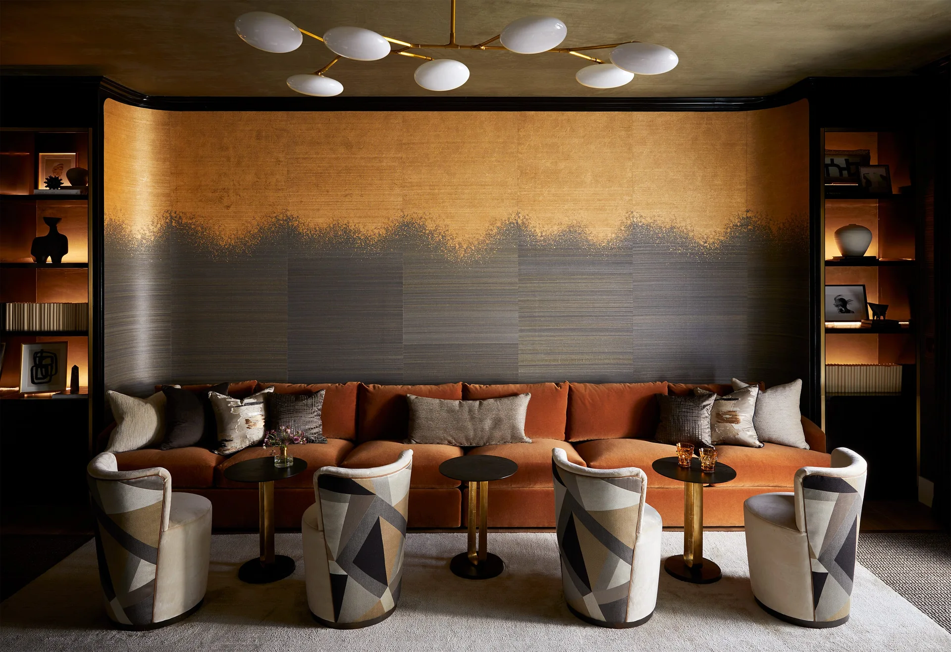 A long orange velvet sofa sits against a curved gold and charcoal metallic wall, flanked by illuminated bookshelves and patterned armchairs.