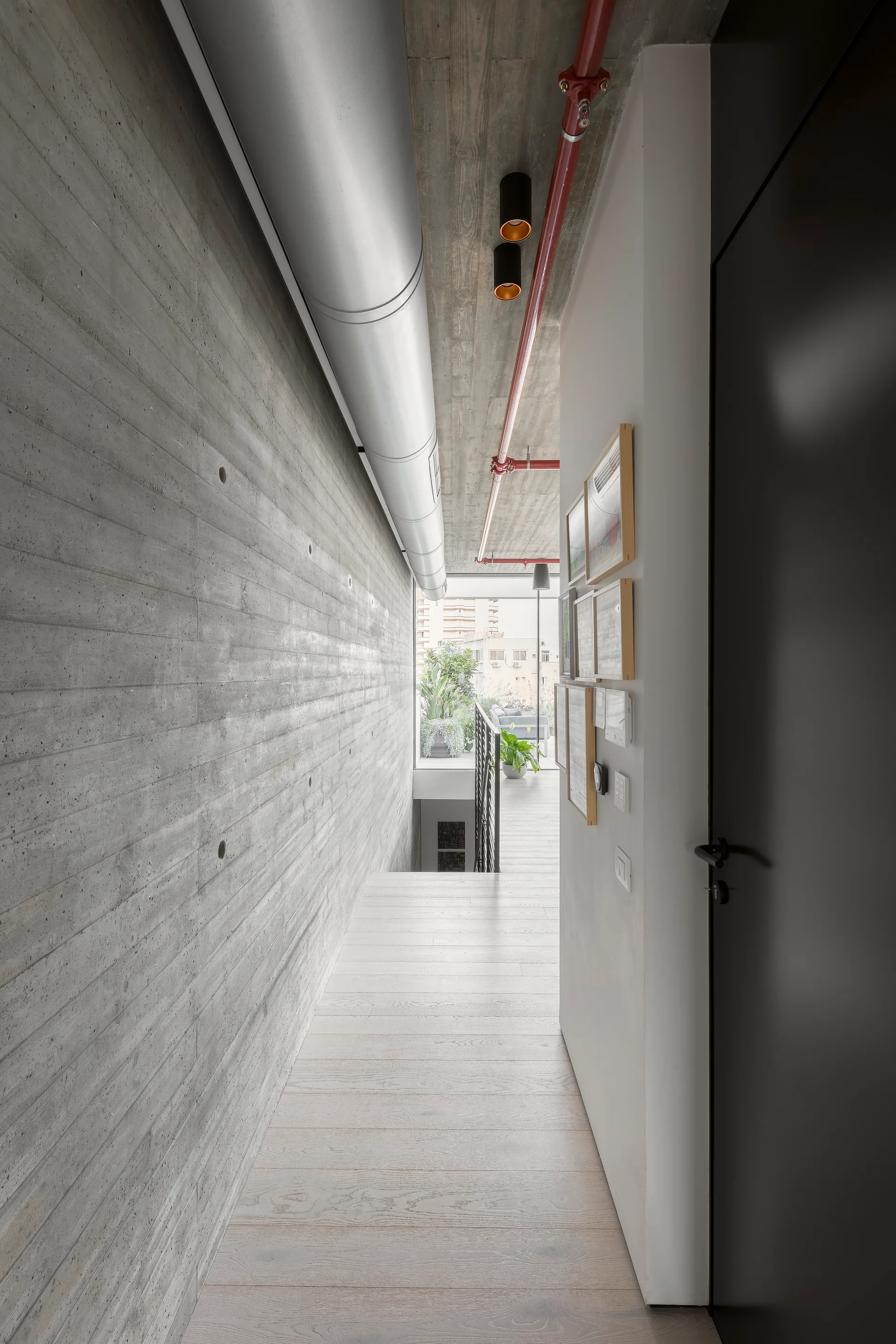 A minimalist industrial hallway with a textured board-formed concrete wall, light wood flooring, and an exposed red pipe on the ceiling.
