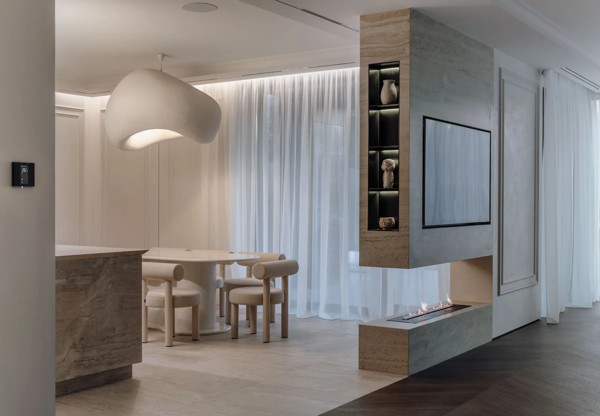 A contemporary dining area featuring an oval table with round-back chairs, a large sculptural pendant light, and a travertine-clad fireplace pillar.