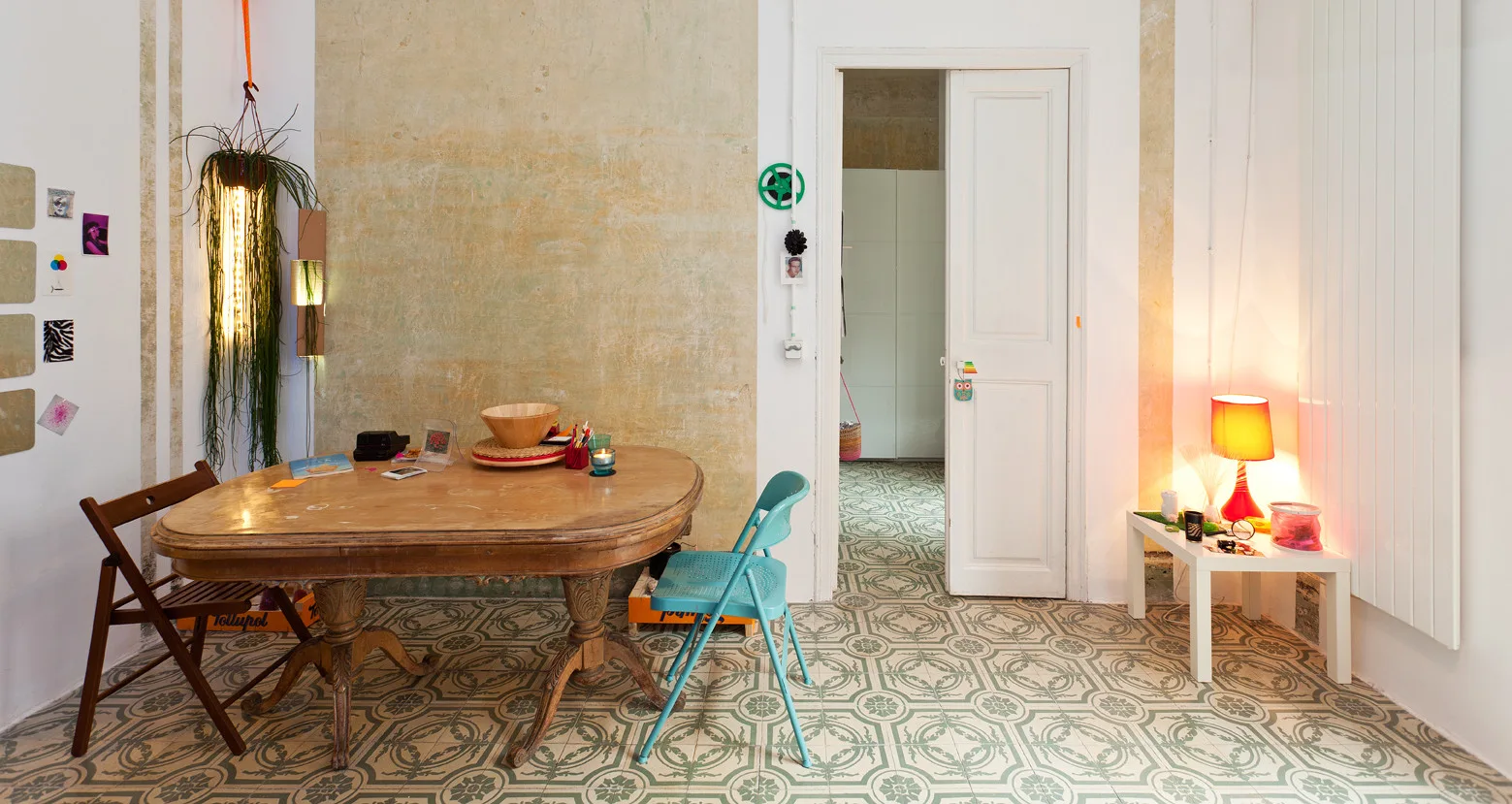 An eclectic dining room featuring an oval wooden table, mismatched chairs, patterned green and white floor tiles, and a distressed plaster wall.