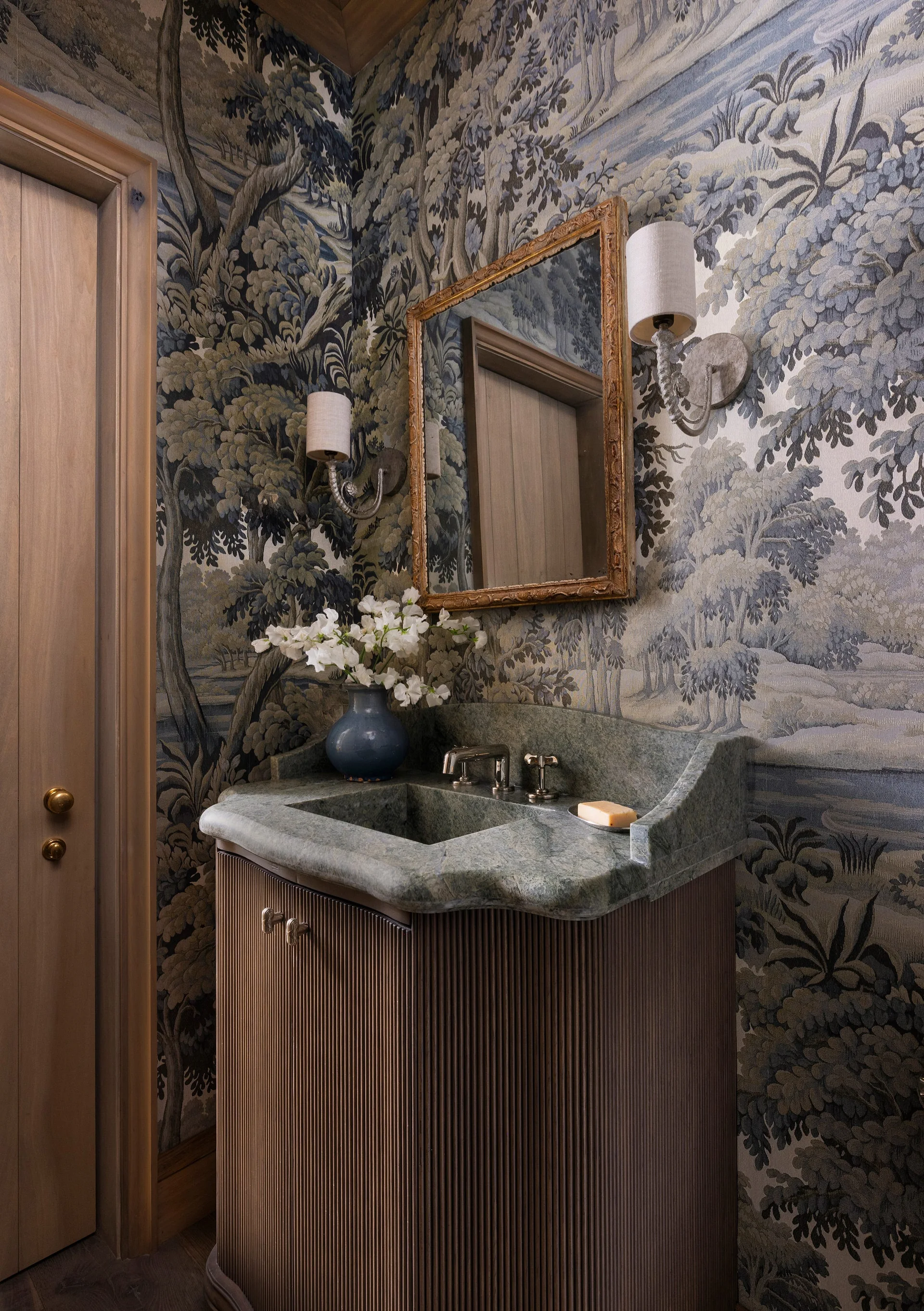 A powder room features a fluted wood vanity with an integrated green stone sink set against scenic landscape wallpaper and a gold-framed mirror.