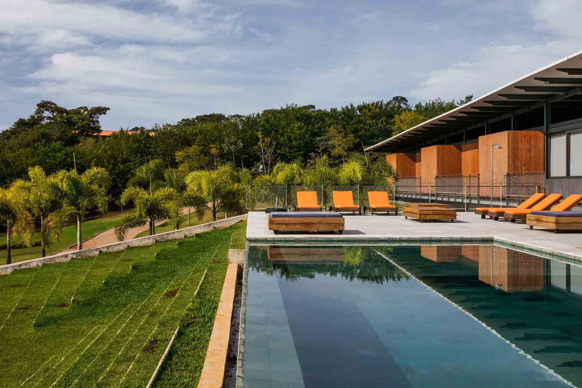 A rectangular swimming pool borders a grey stone patio featuring orange lounge chairs, wooden daybeds, and a sloped green lawn with palm trees.