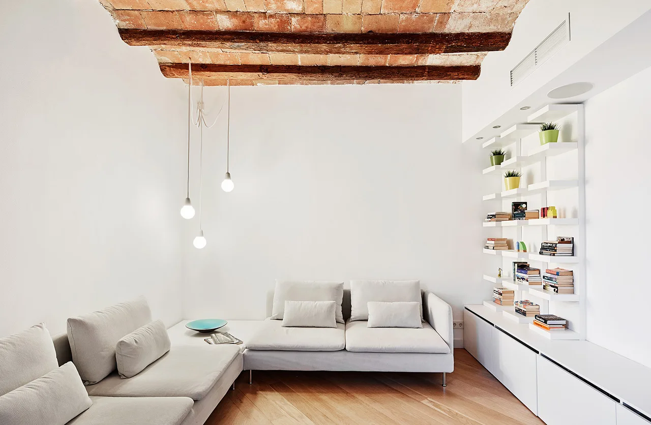 A minimalist white living room features a sectional sofa, white wall shelving, and a vaulted brick ceiling with a dark wooden beam.