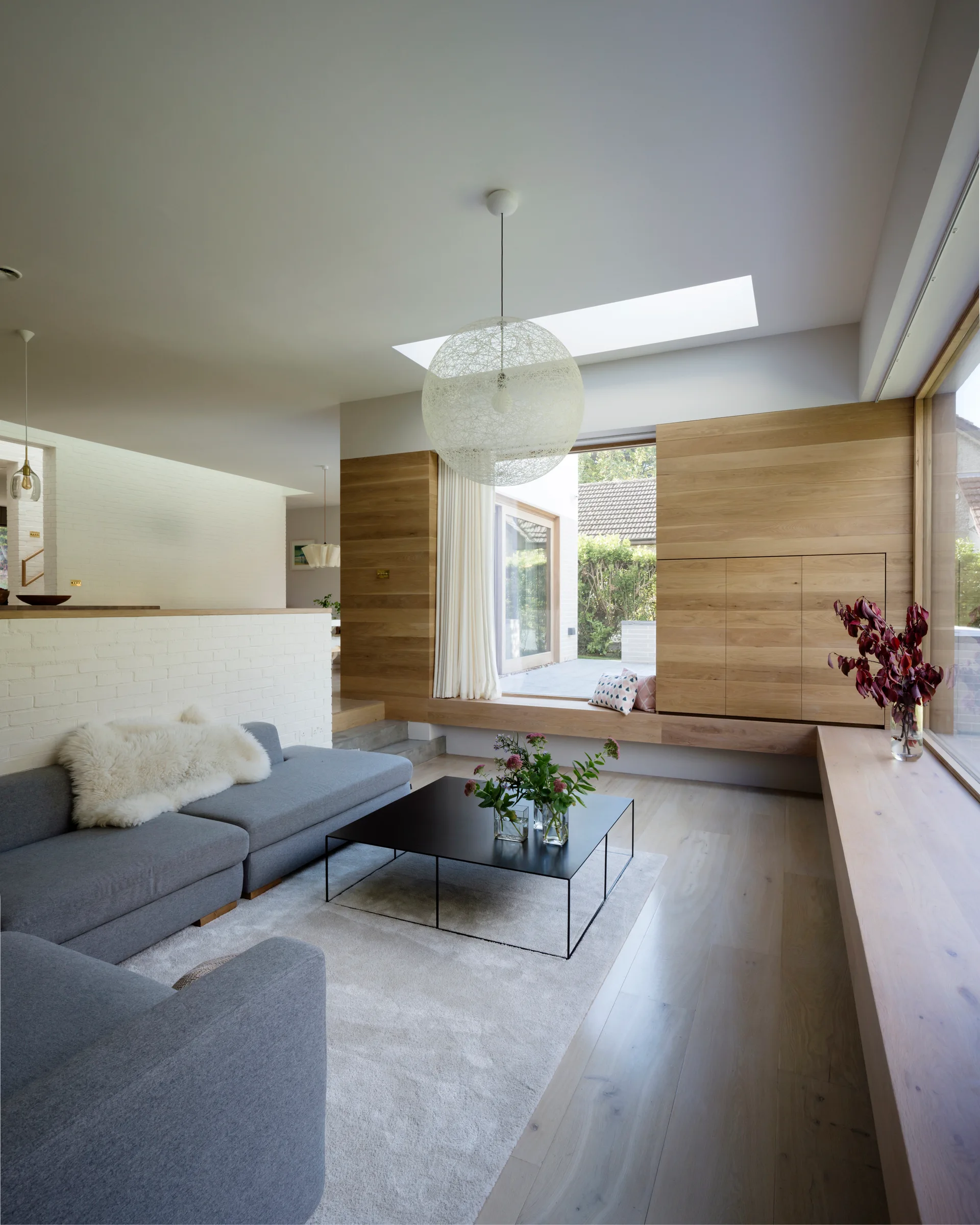 A modern living room features a grey sectional sofa, black coffee table, and an L-shaped light wood window seat under a skylight.