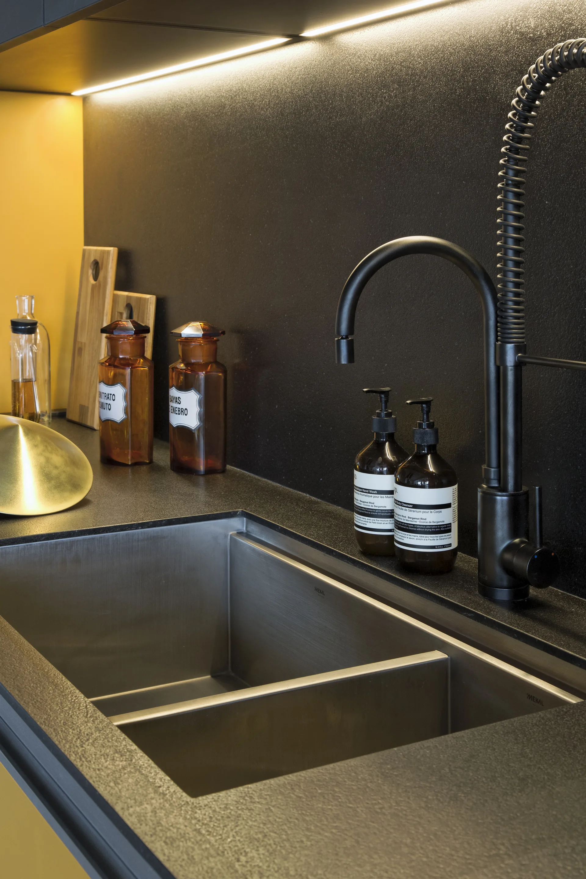 A stainless steel undermount kitchen sink set into a dark matte countertop with a black pull-down coil faucet, amber bottles, and under-cabinet strip lighting.