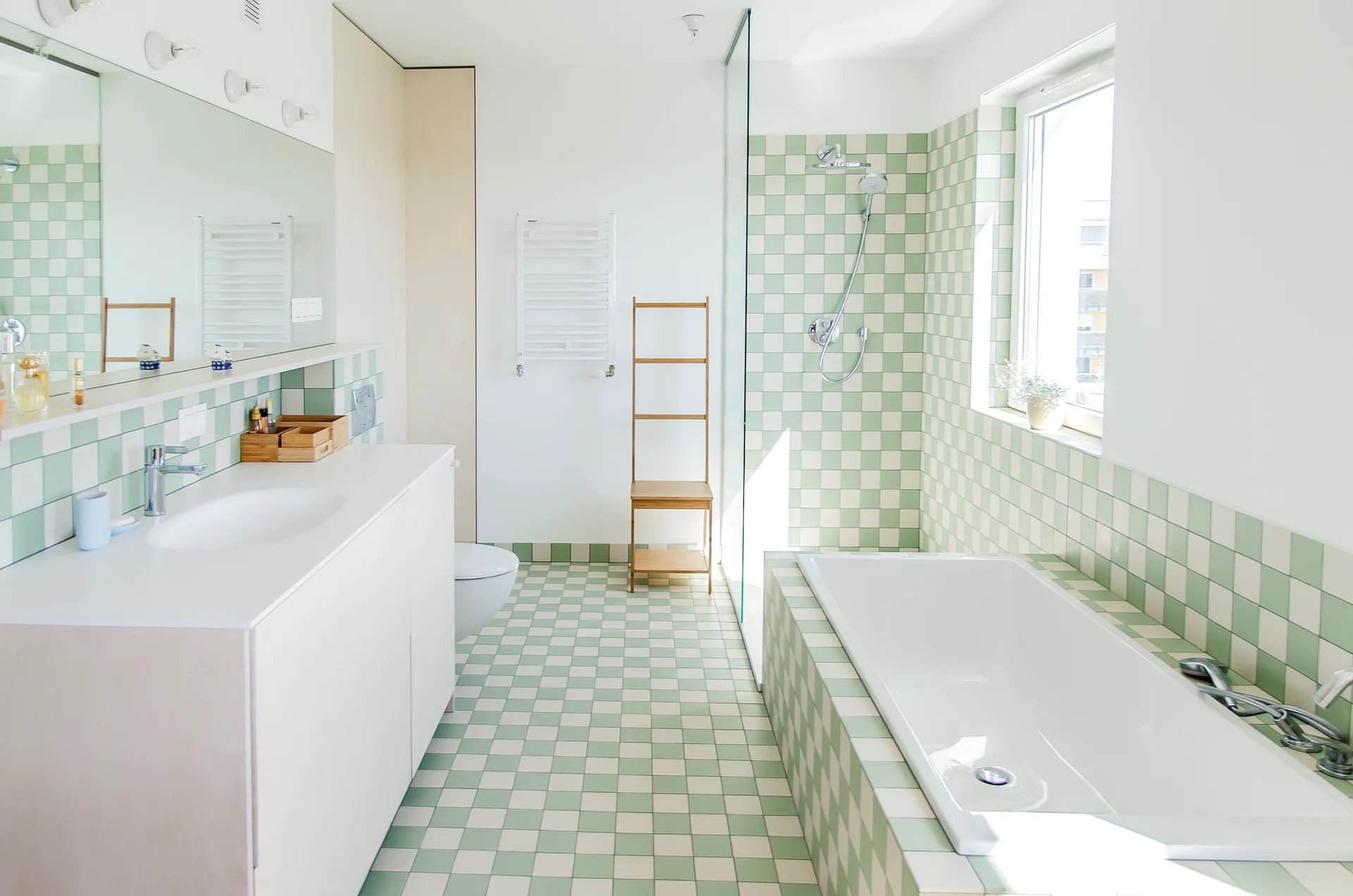 A modern bathroom featuring sage green and white checkered tiles on the floor and bathtub surround, alongside a long white vanity and wall mirror.