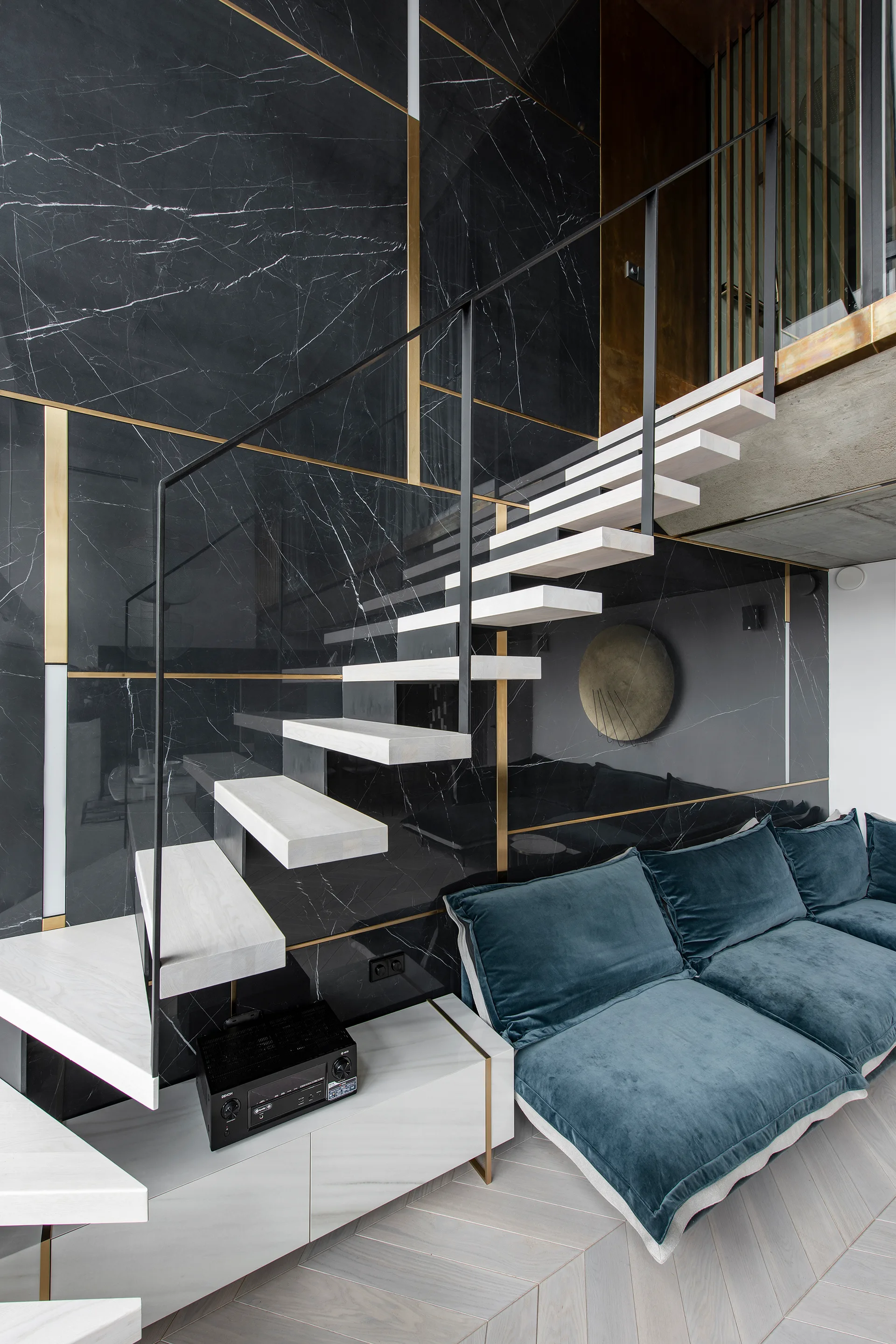 A modern space with a floating light wood staircase against a black marble wall with brass accents next to a teal velvet sofa.
