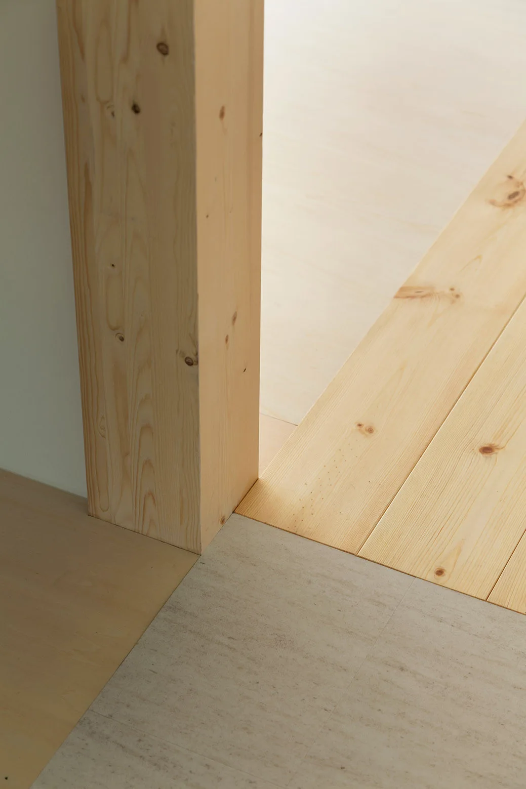 A close-up architectural view showing a light wood post meeting a transition between wooden planks and beige stone-textured tile floors.