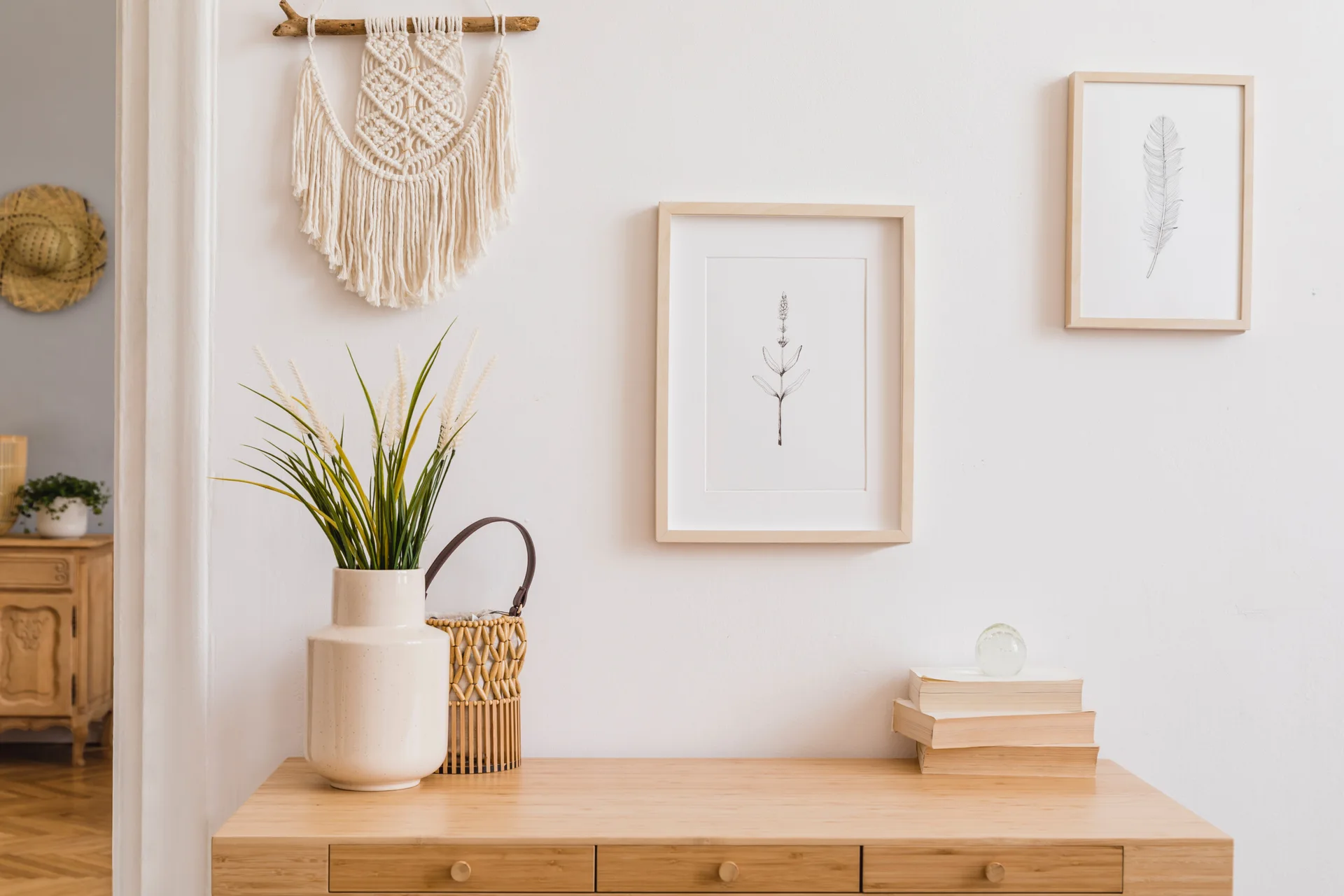 A light wood console table styled with a white vase, books, and a woven basket against a white wall with framed botanical prints and a macrame hanging.