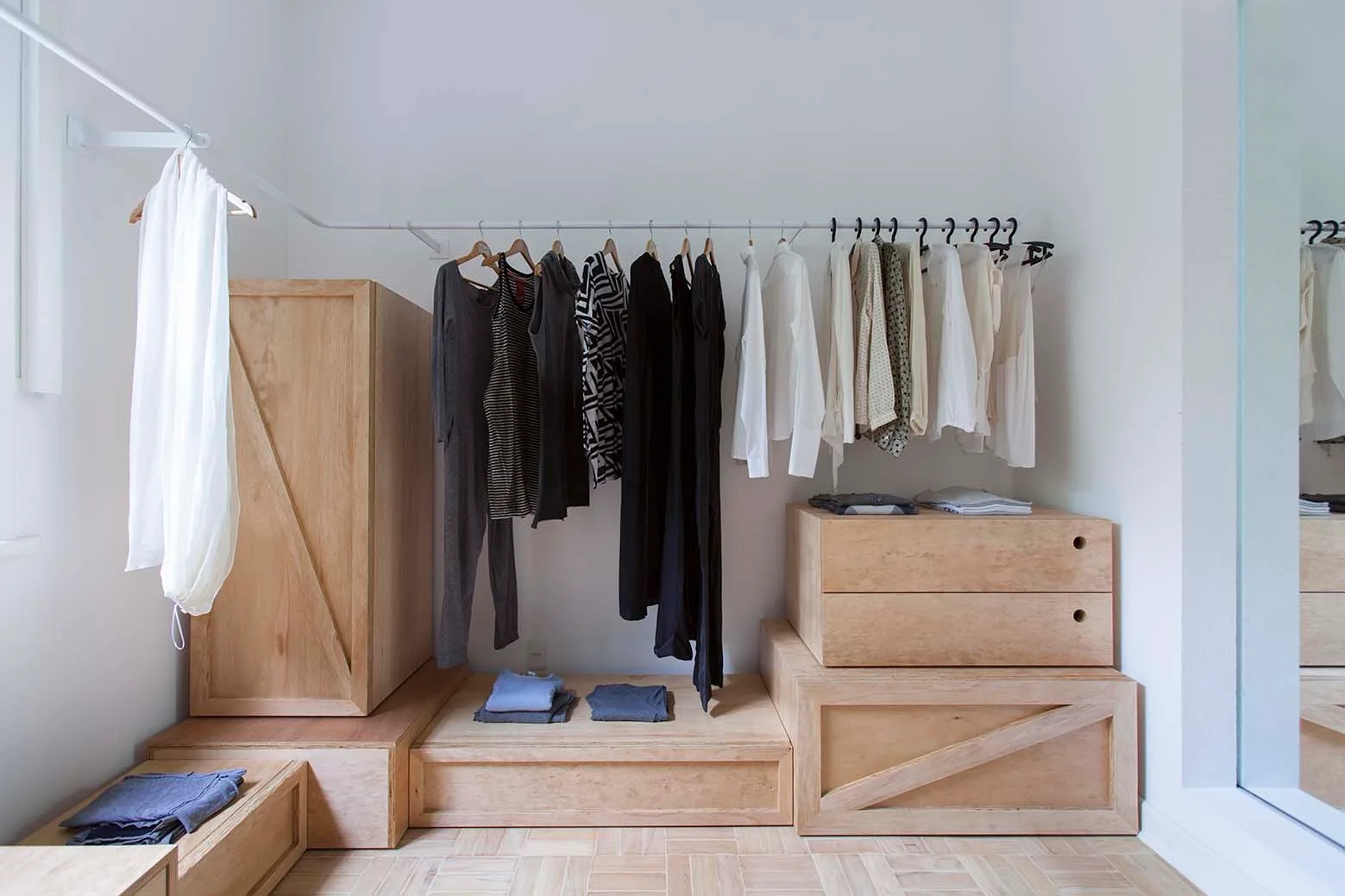 A minimalist closet features light wood modular storage crates and drawers beneath a white wall-mounted clothing rail on a parquet floor.