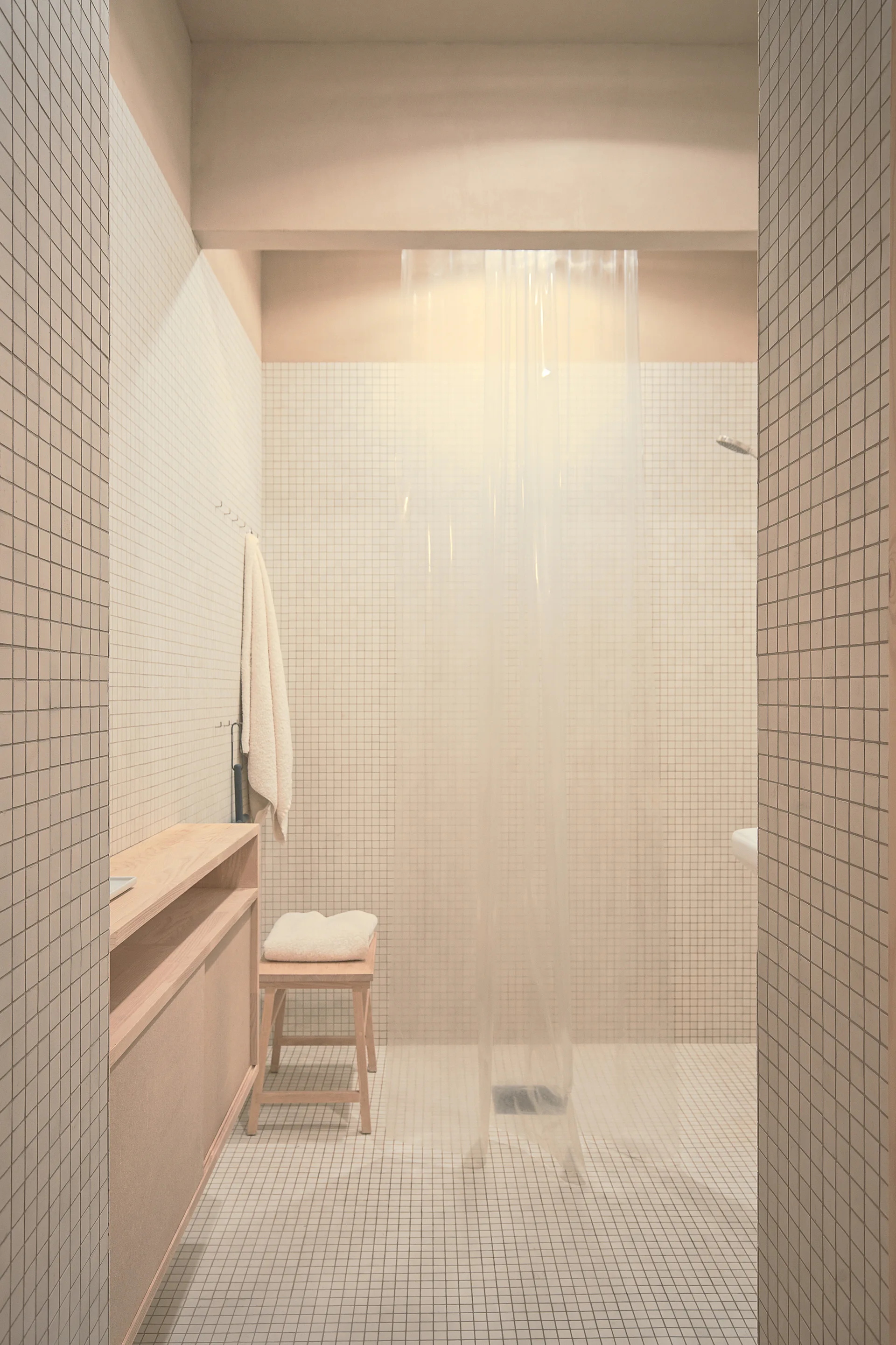 A minimalist beige bathroom features small square grid tiles on the walls and floor, a light wood vanity, a matching stool, and a walk-in shower.