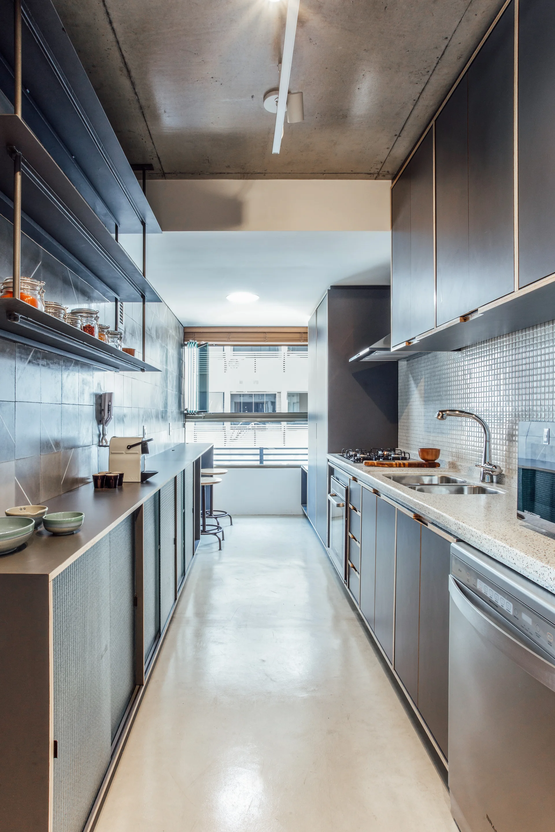 A narrow galley kitchen with dark grey cabinets, a polished light floor, a raw concrete ceiling, and a window at the far end.