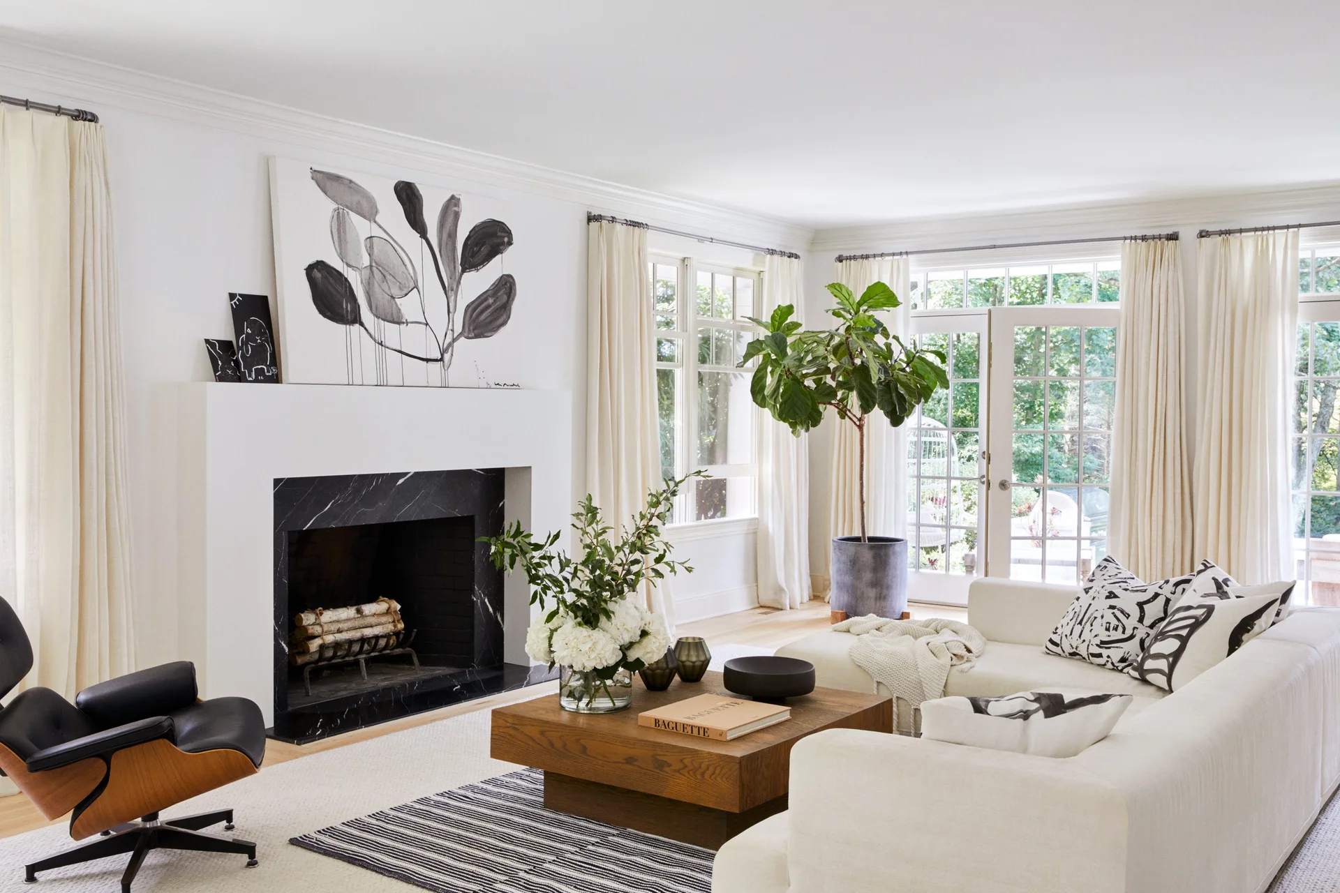 A bright living room featuring a white sectional sofa, a black marble fireplace, layered area rugs, and a tall potted fiddle leaf fig tree.