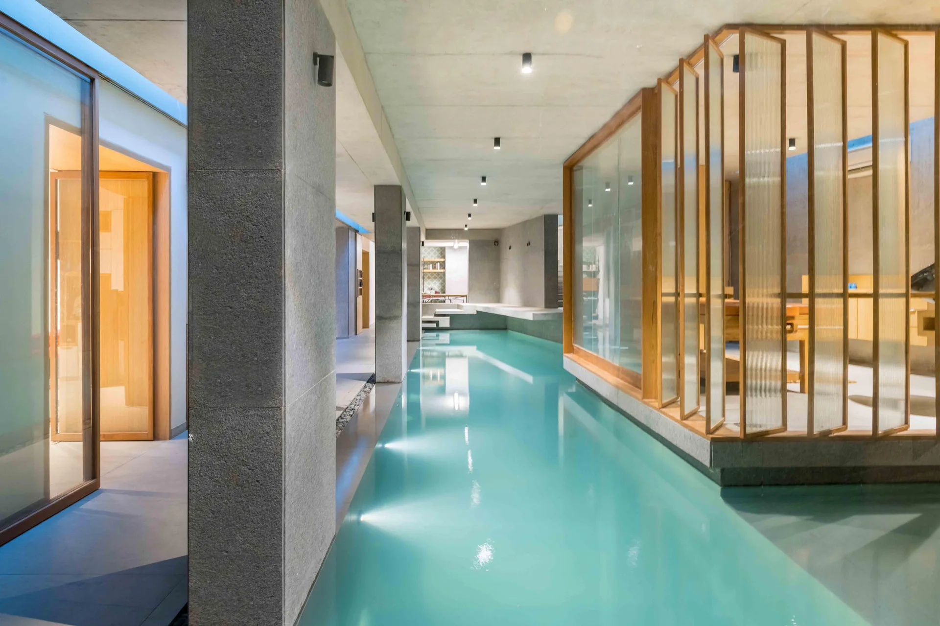 An indoor lap pool with turquoise water is bordered by concrete columns and a wood-framed fluted glass partition.