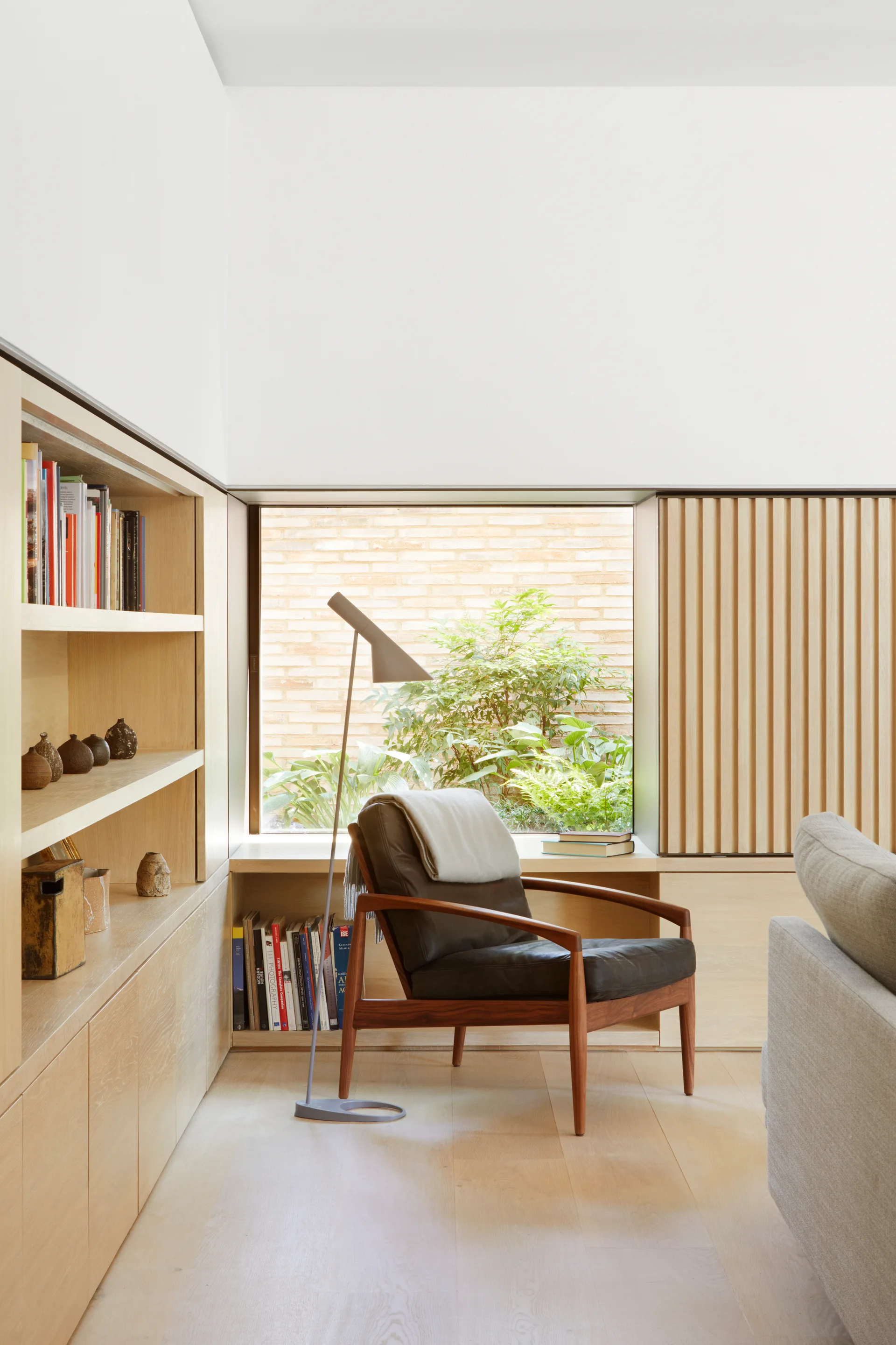 A leather armchair and slim metal floor lamp sit in a light wood reading nook next to built-in shelving and a large window.
