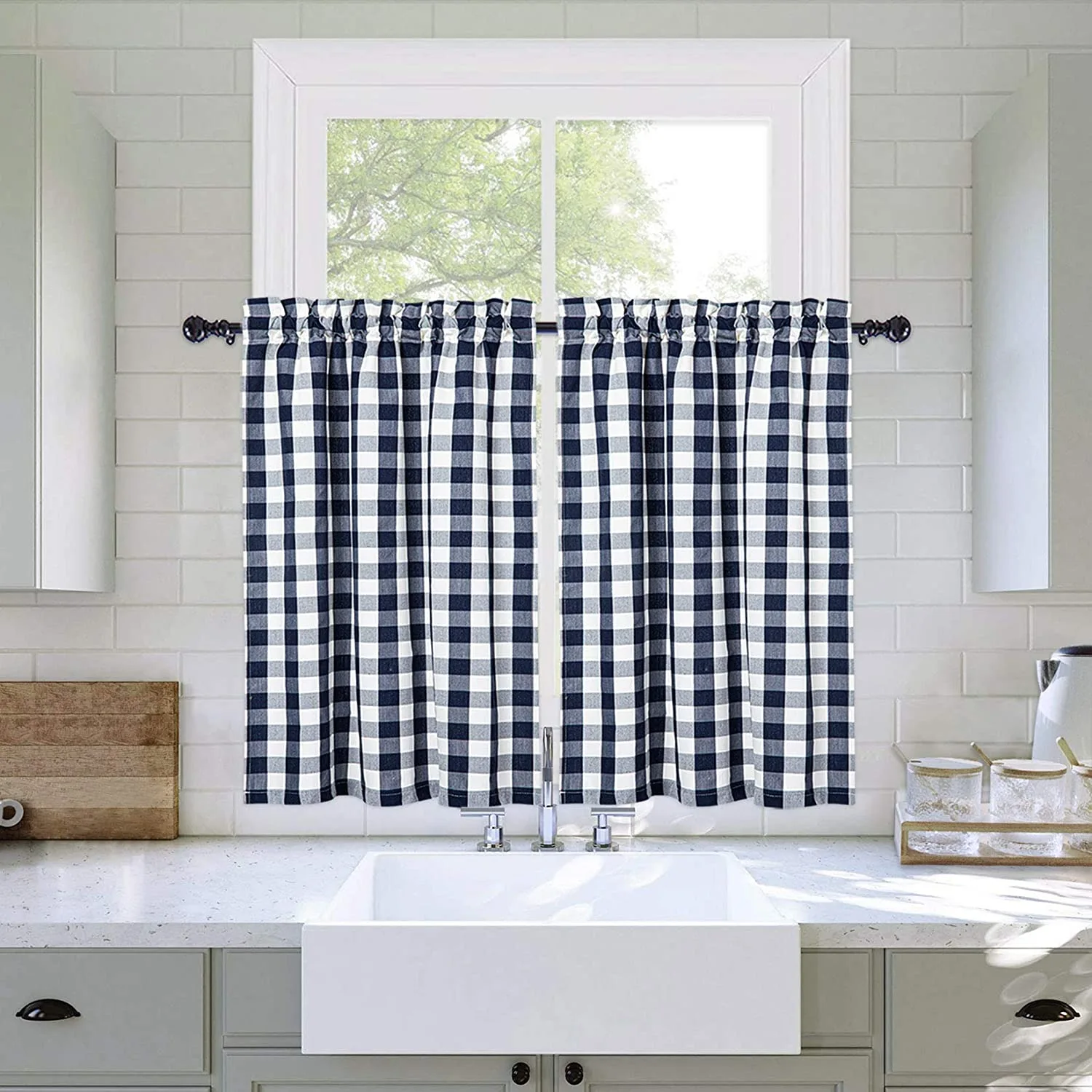 A white farmhouse kitchen featuring a white apron sink beneath a window decorated with navy and white buffalo check cafe curtains.