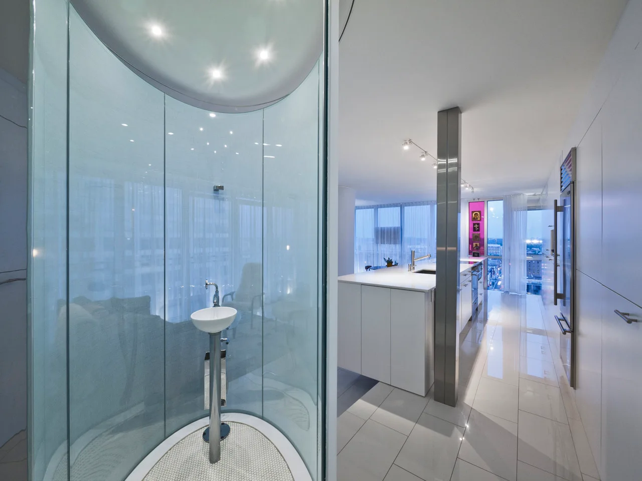 A modern apartment kitchen with glossy white floor tiles sits adjacent to a transparent curved glass shower stall containing a pedestal sink.