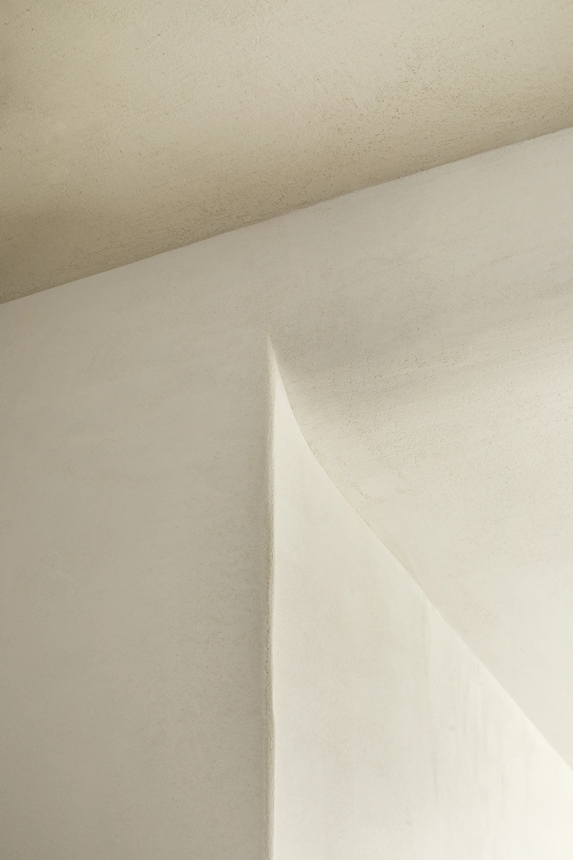 A close-up view of a minimalist architectural corner featuring smooth, matte cream plaster walls with a sharp vertical edge and a curved transition.