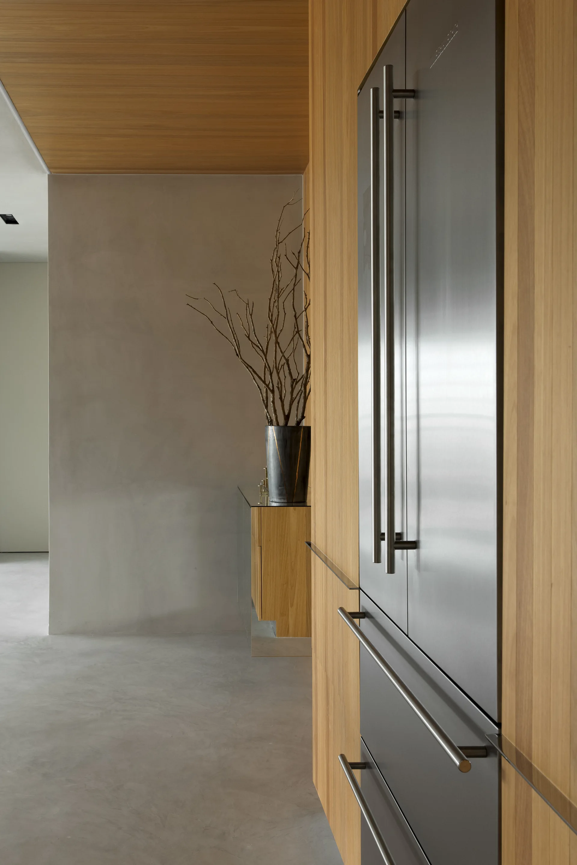An integrated stainless steel refrigerator is built into warm wood wall panels next to a smooth grey concrete floor and a plaster wall.