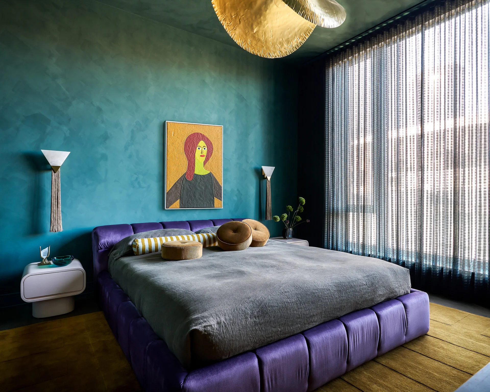 A modern bedroom features a low purple upholstered bed against a textured teal wall with a yellow portrait painting and a large gold pendant light.