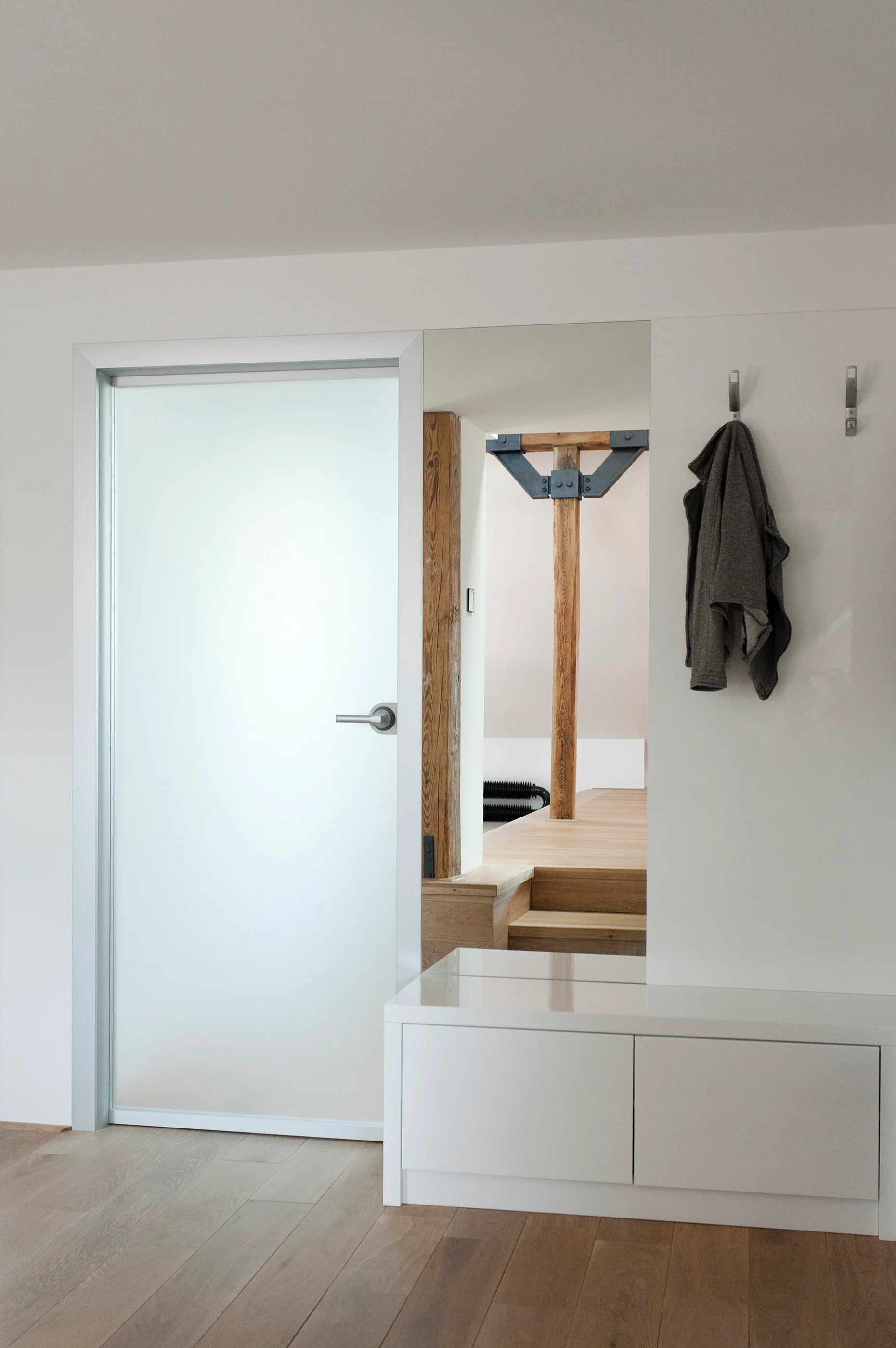 A minimalist entryway features a frosted glass door, a full-length wall mirror, a white storage bench, and metal coat hooks on a white wall.