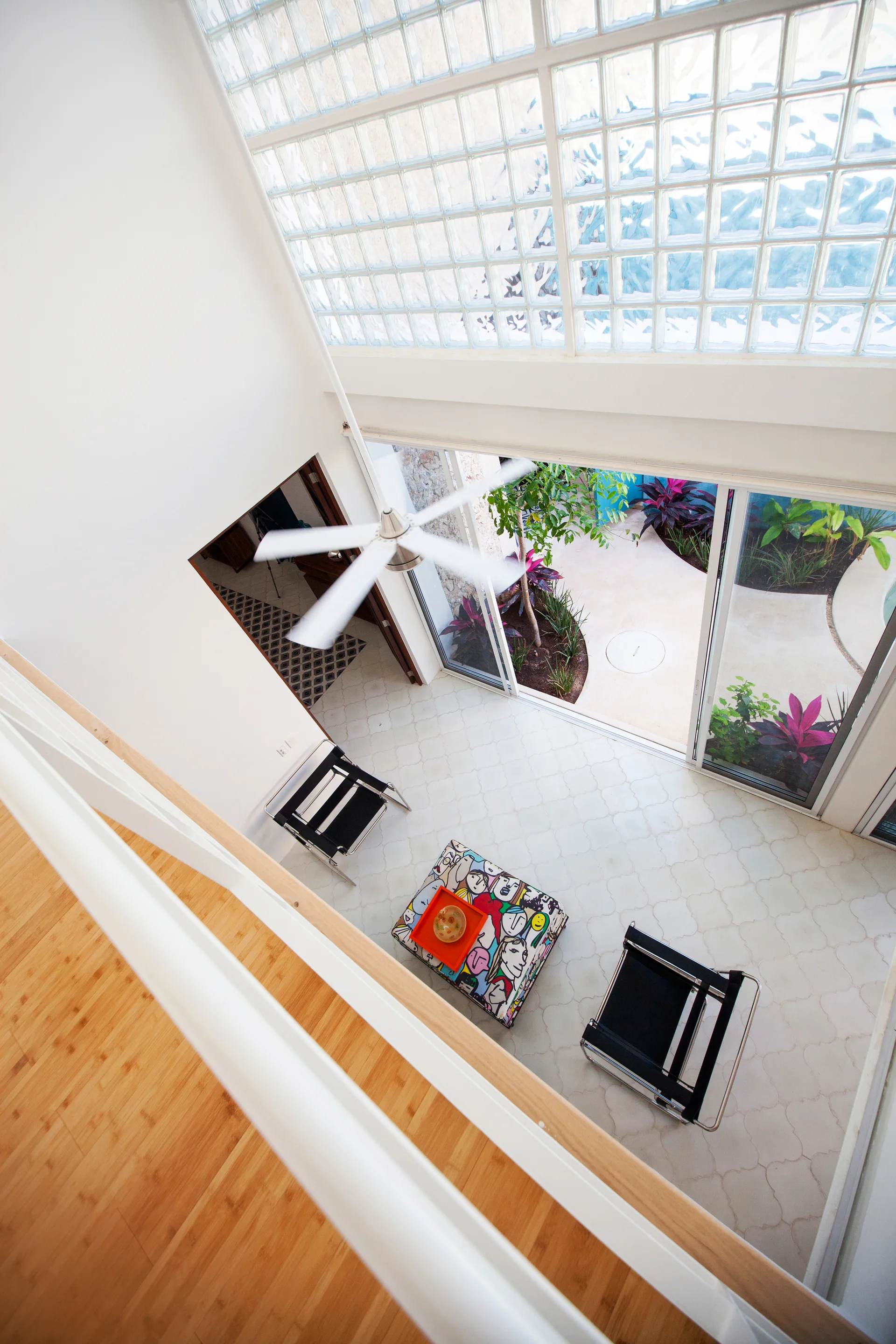 A high-angle view of a double-height white living room with black steel armchairs, a patterned ottoman, and glass sliding doors leading to a courtyard.