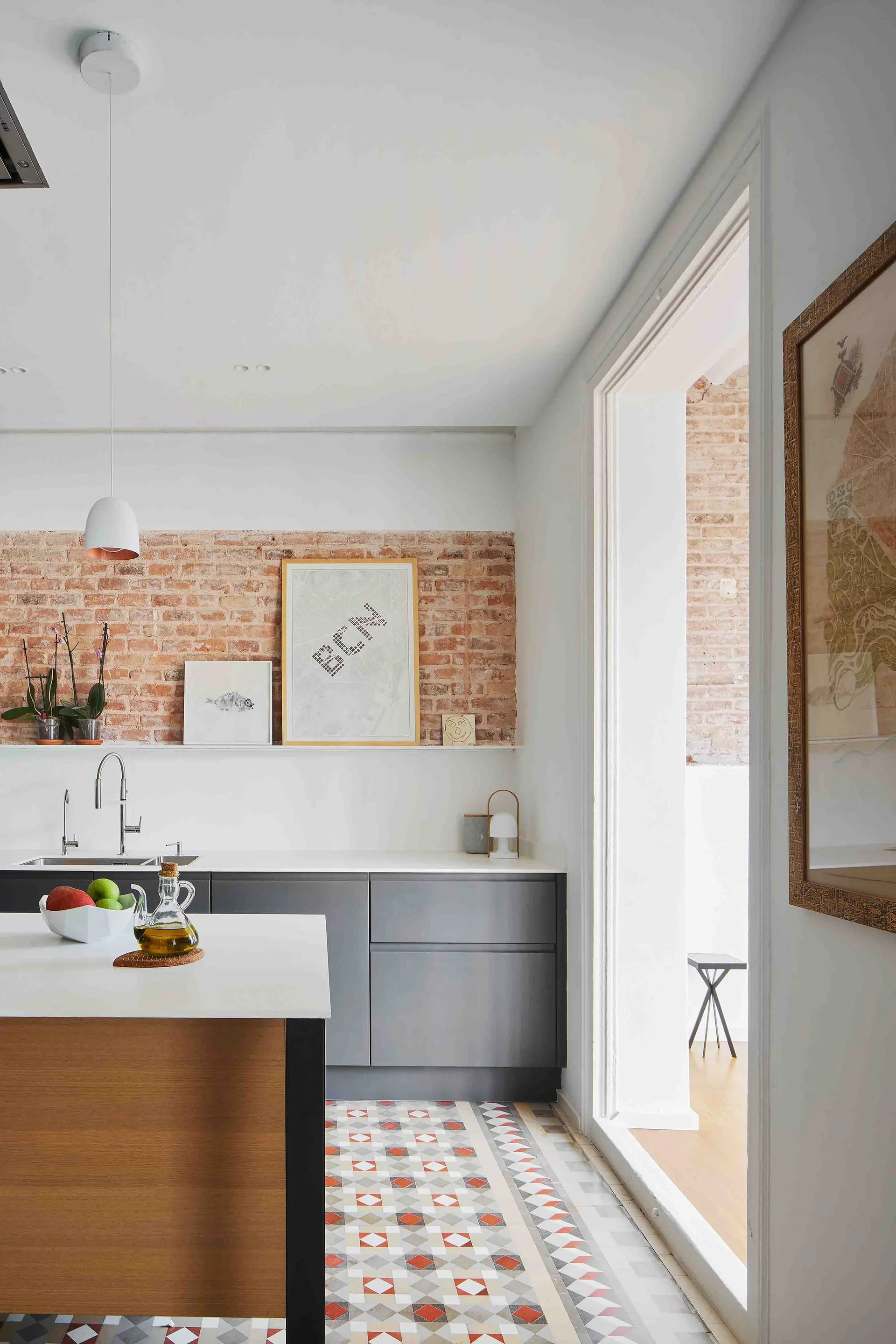 A modern kitchen with a wood-based island, dark grey cabinets, patterned geometric floor tiles, and an exposed red brick wall.