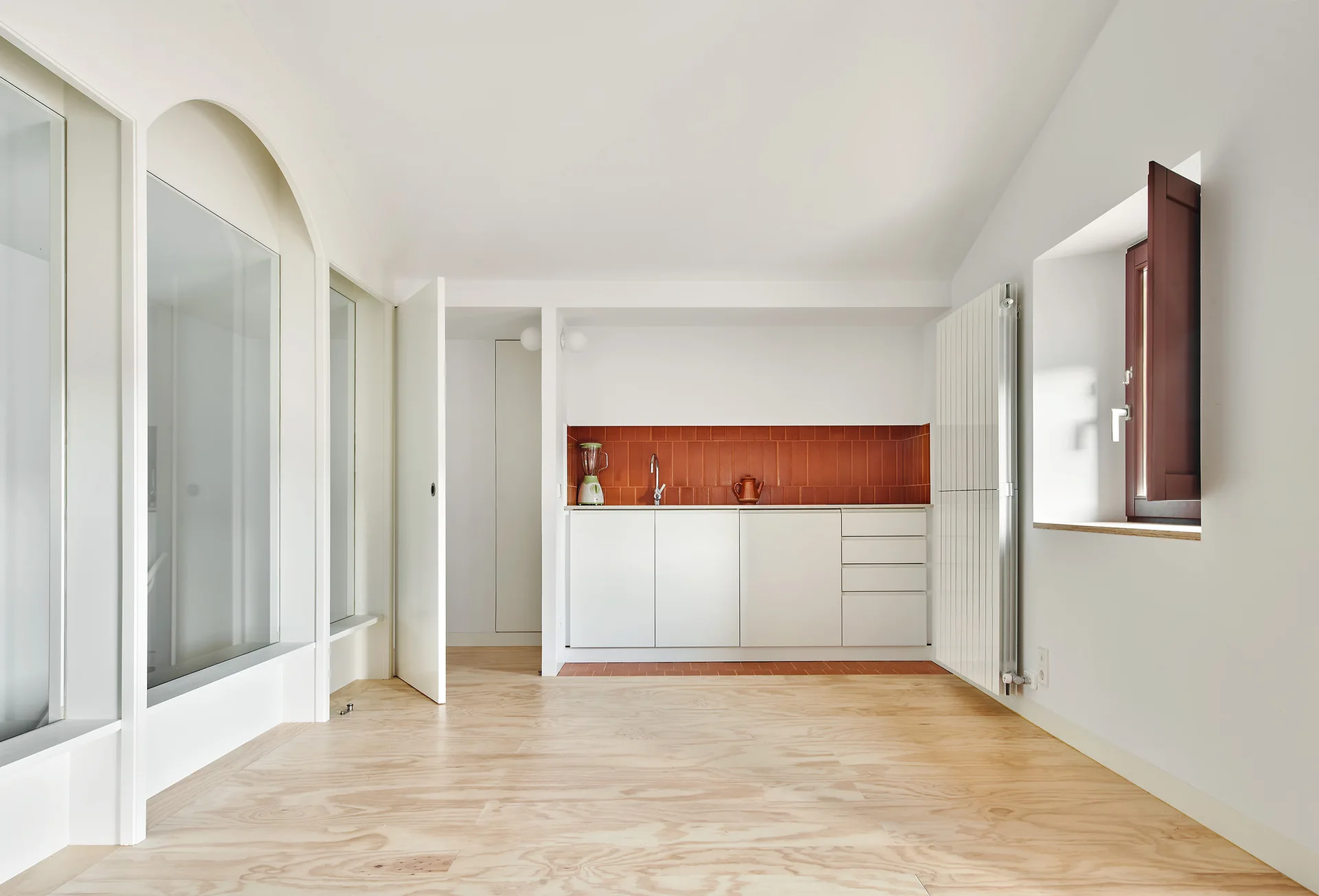 A minimalist kitchen featuring white flat-panel cabinets, a vertical terracotta tile backsplash, light wood floors, and arched glass partitions on the left.