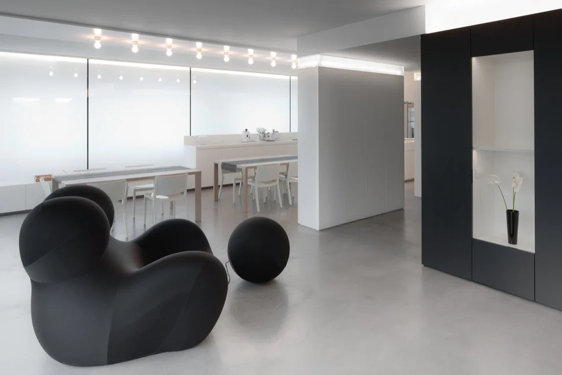 A sculptural black armchair sits in a minimalist grey and white room featuring built-in black cabinetry and a distant dining area.