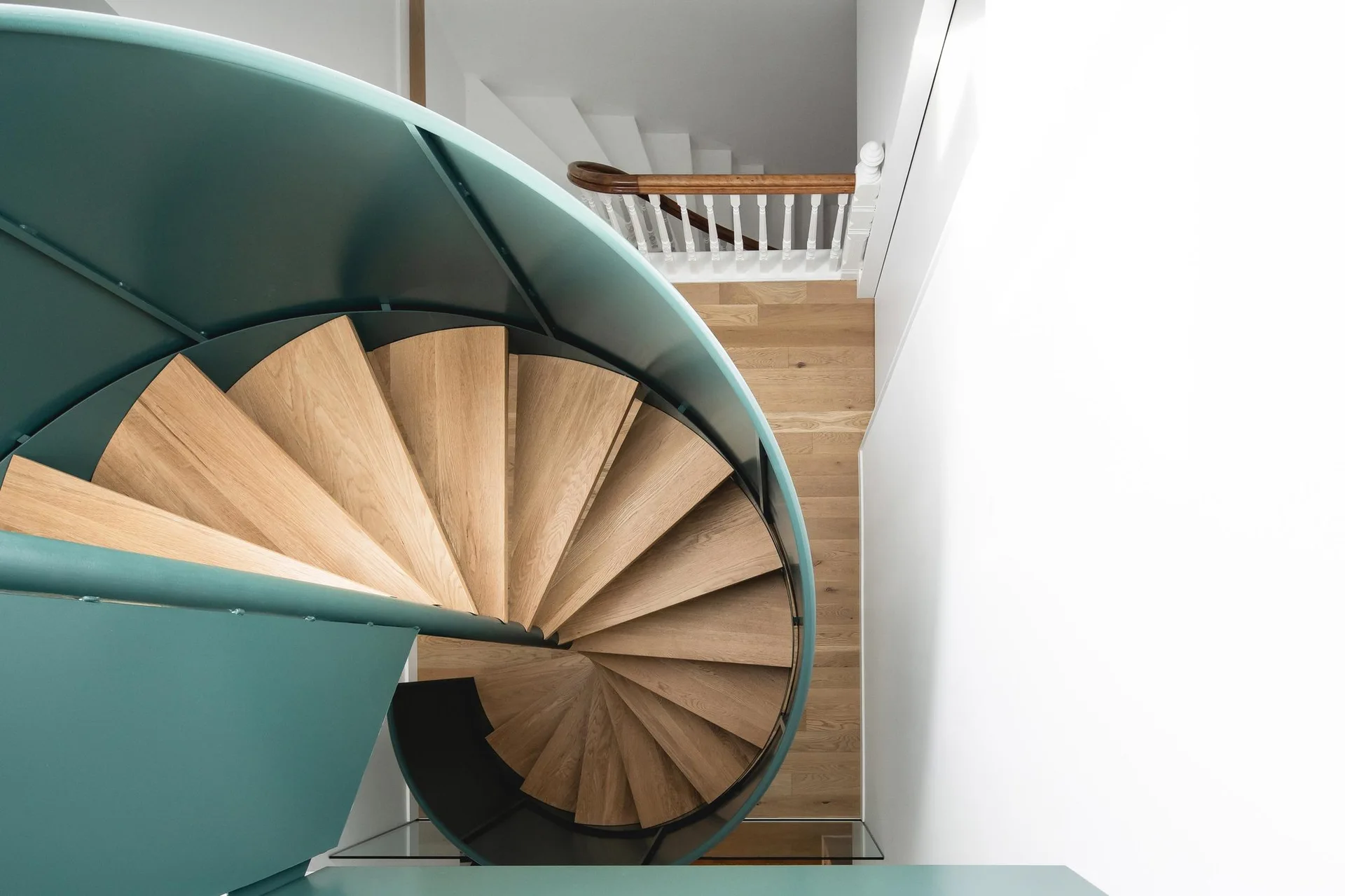 Looking down a spiral staircase featuring light oak wood treads surrounded by a curved teal metal railing against white walls.