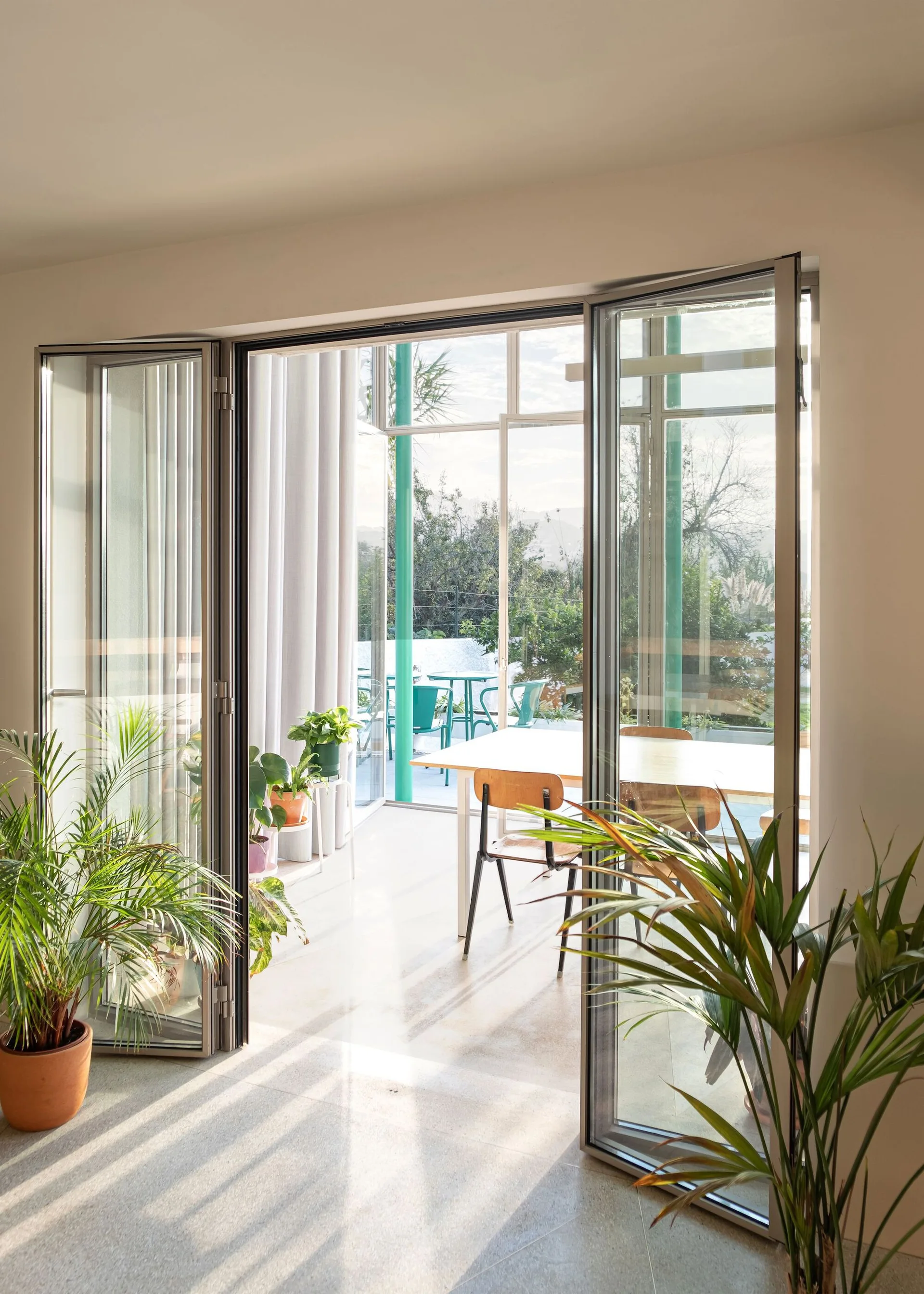 Large metal-framed bifold doors open from a light gray tiled room to a sunny outdoor terrace with a dining table and potted plants.