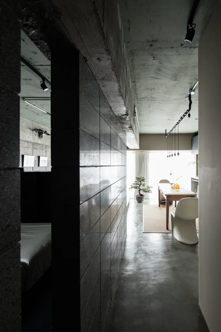 A dark corridor with a glossy concrete block wall leads to a bright dining space with a wooden table, white chairs, and an exposed concrete ceiling.