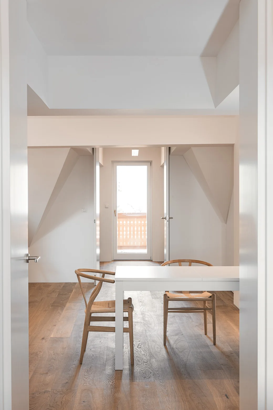 A white dining table and two wooden wishbone chairs sit on light wood flooring in a minimalist room with white angled walls and a glass balcony door.