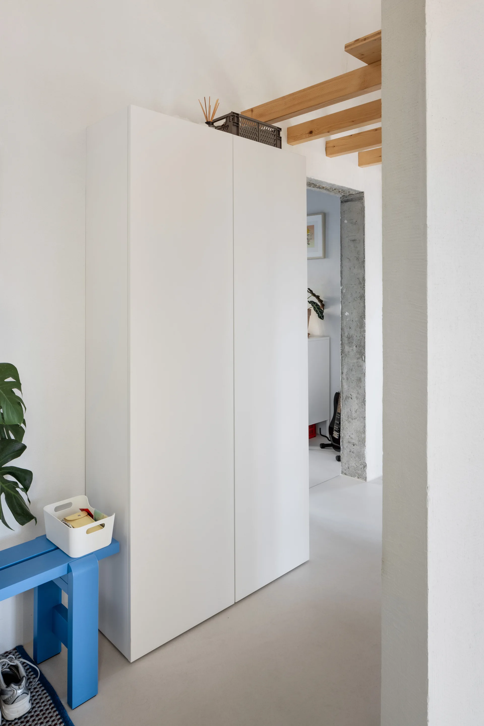 A minimalist white entryway featuring a tall white wardrobe, a bright blue wooden bench, and an exposed raw concrete doorway.