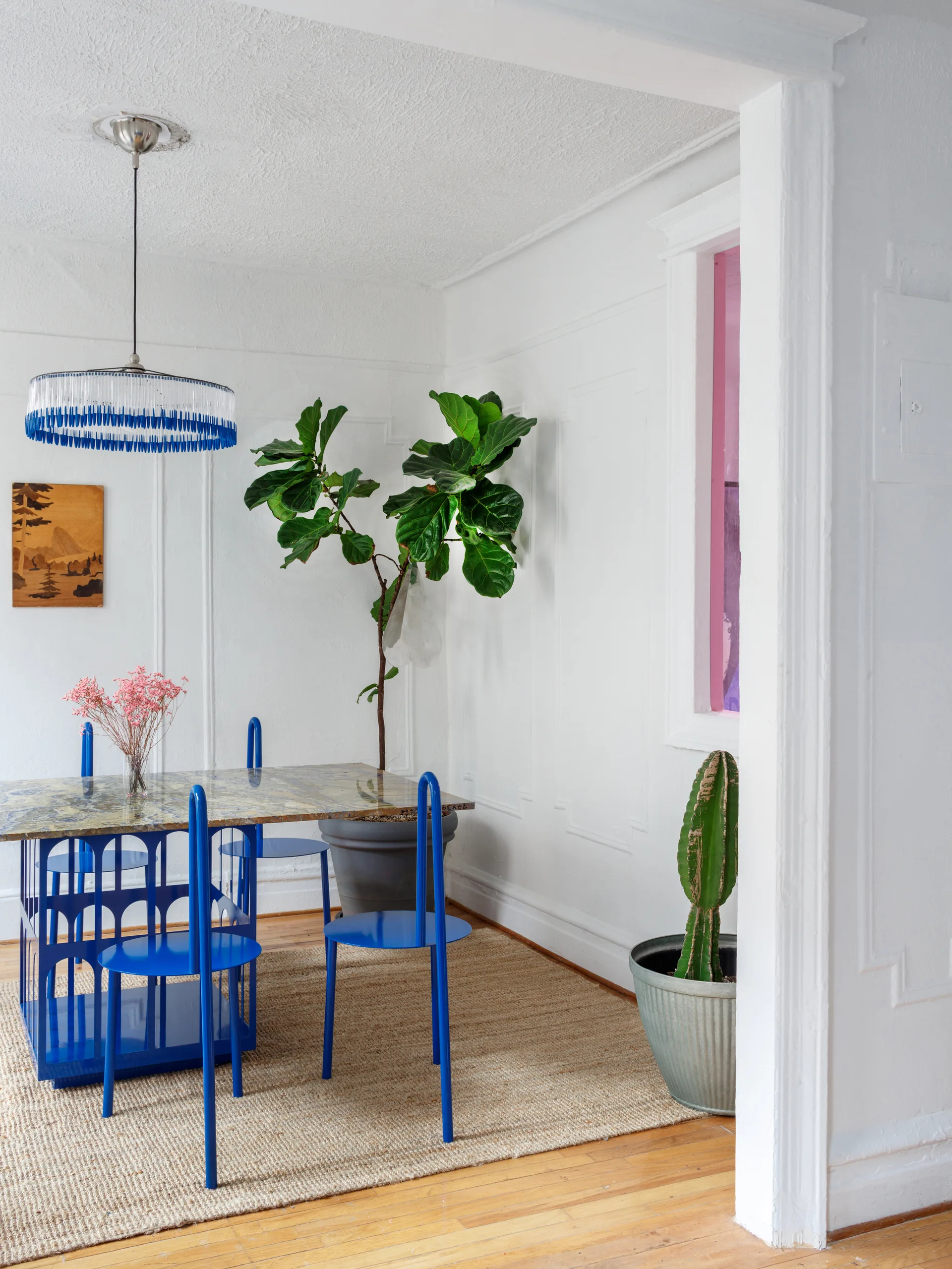Cobalt blue metal chairs surround a marble-topped table on a jute rug in a white dining room with paneled walls and potted plants.