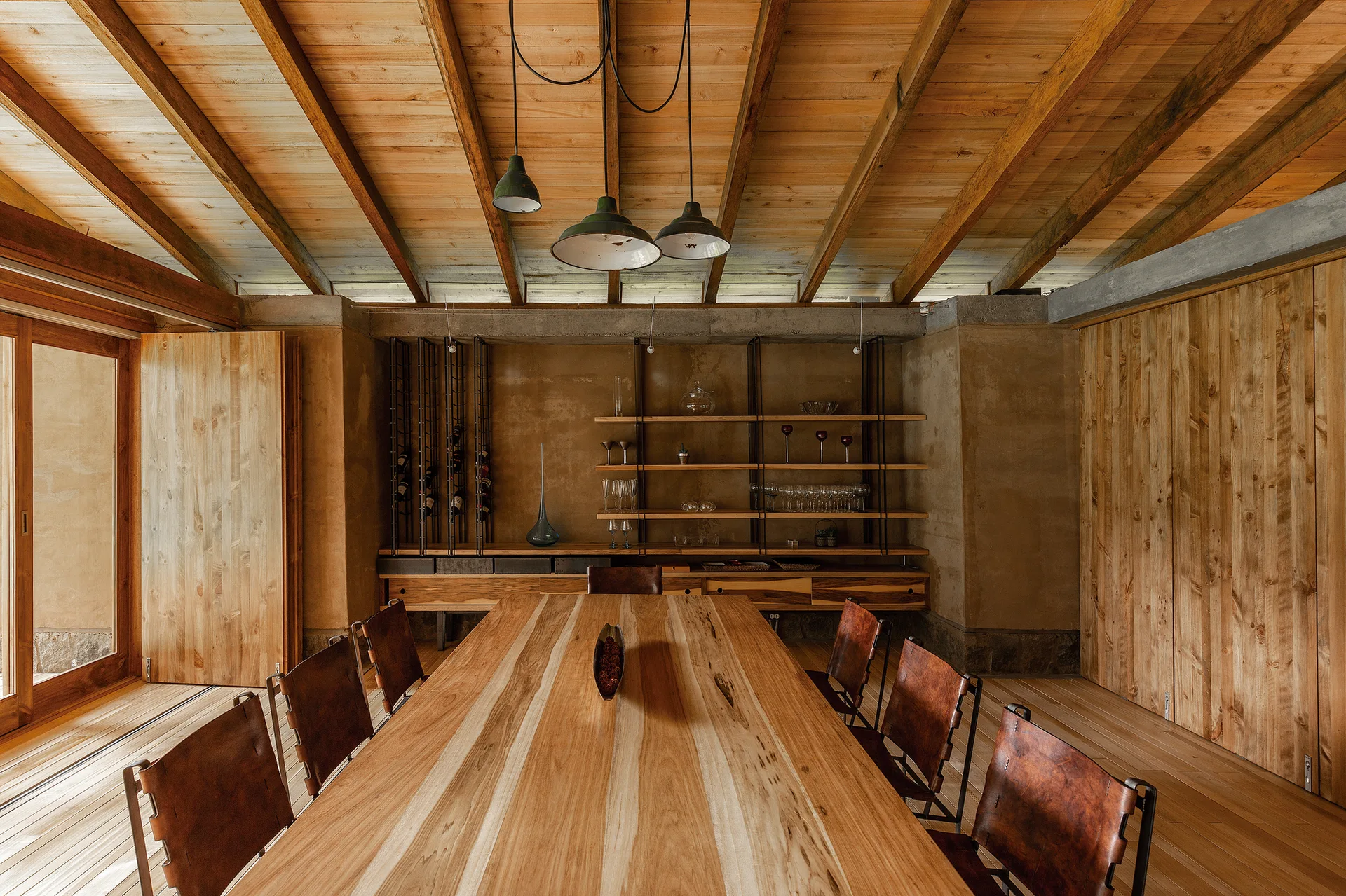 A rustic dining room features a long wooden table surrounded by brown leather chairs under an exposed timber ceiling with three pendant lights.