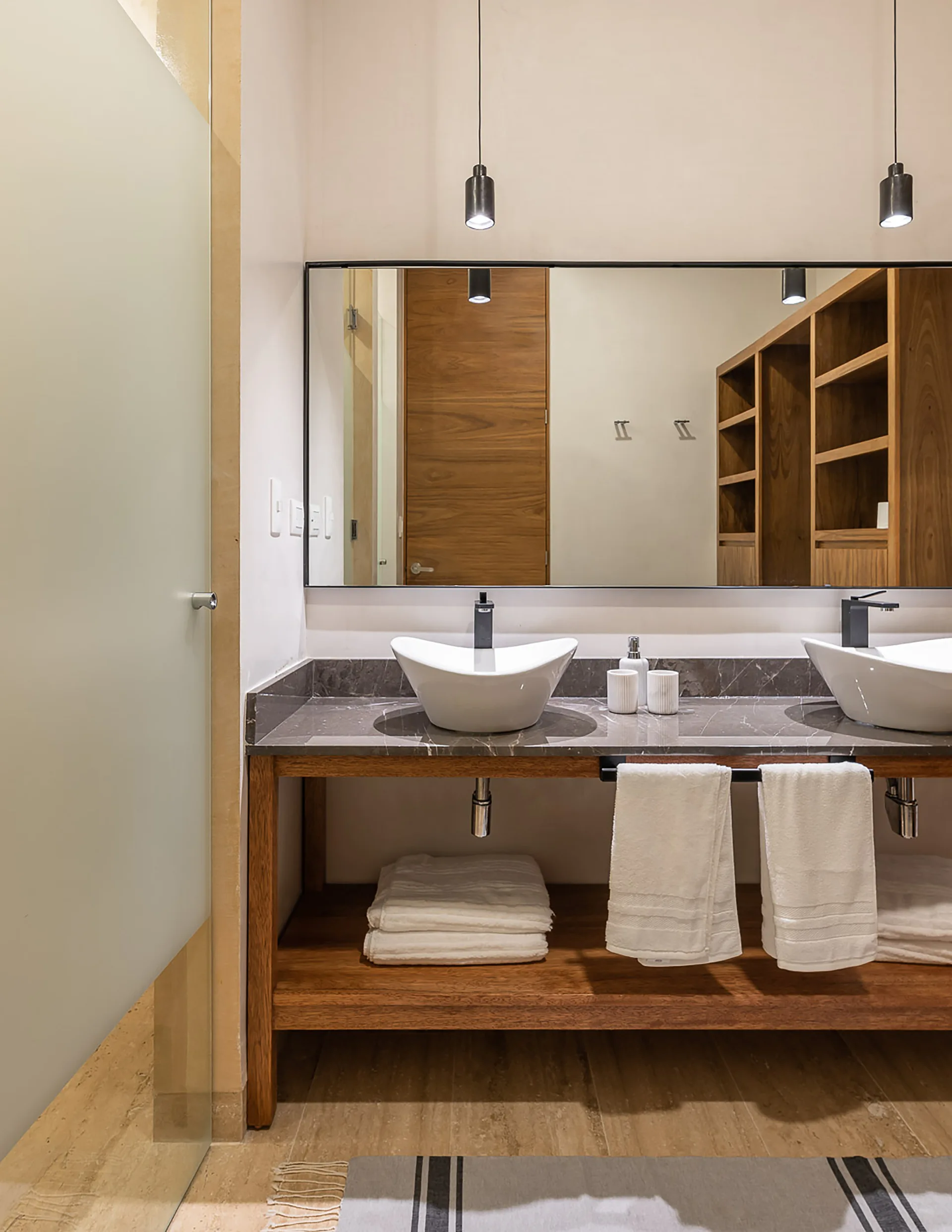 A modern double bathroom vanity features two white vessel sinks on a grey stone countertop with a natural wood base and open shelving.