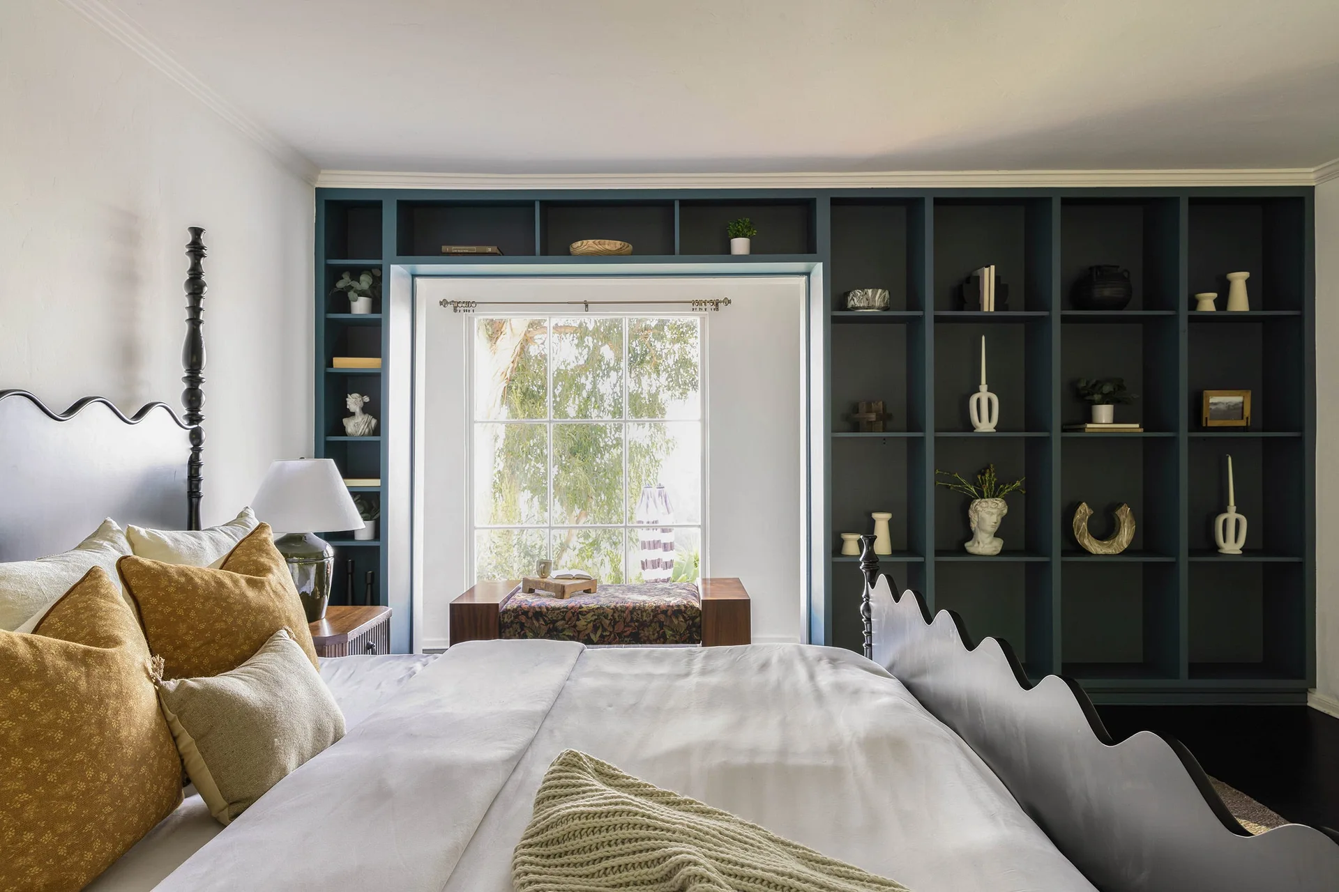 A bedroom features a bed with a scalloped black headboard next to a deep blue built-in shelving unit surrounding a window.