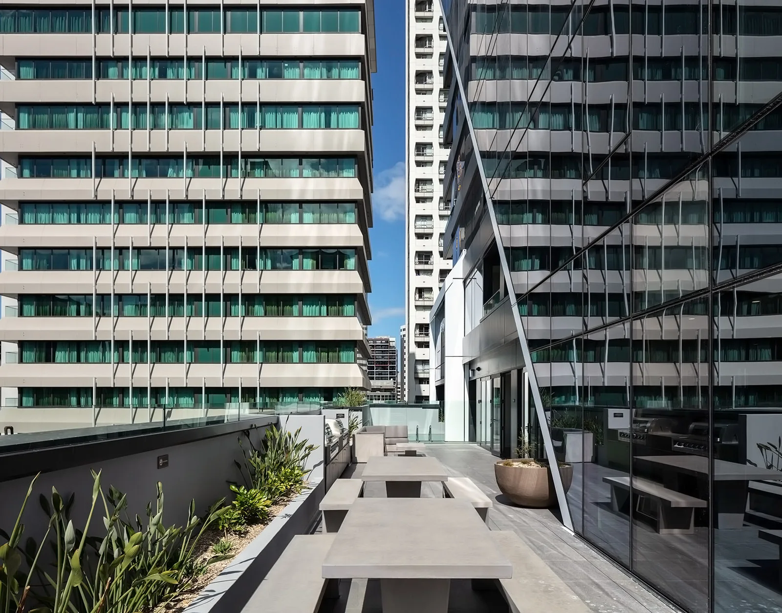Grey concrete picnic tables sit on a modern urban terrace next to a reflective glass building facade and small planters.